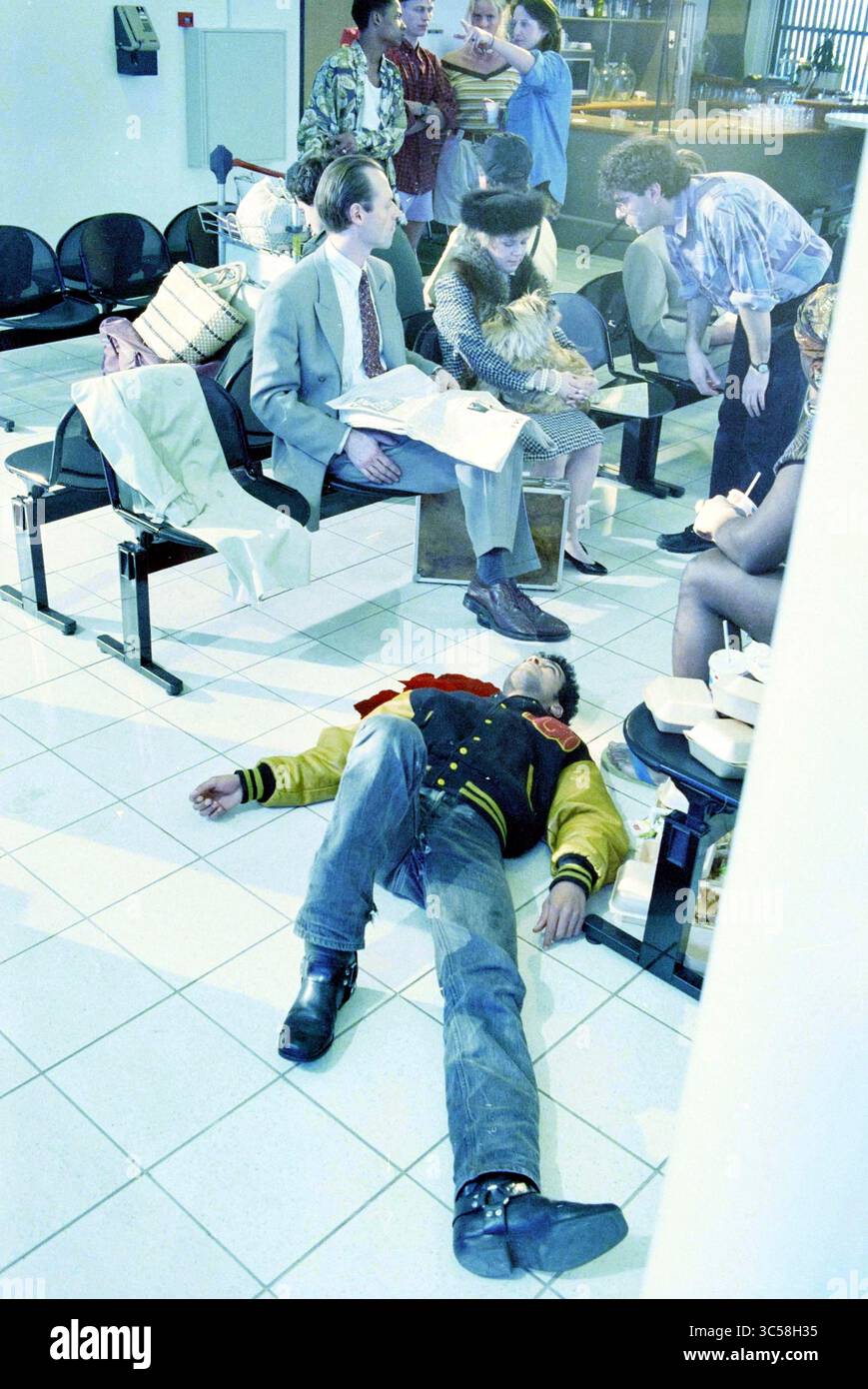 Registrazione film Felisan terminal, IJm., IJmuiden, Paesi Bassi, 22-03-1995 Whizgle News, Dutch Desk, Paesi Bassi, 1950-2000 in una vivace area d'attesa, un gruppo eterogeneo di persone è impegnato in varie attività, mentre una persona è sdraiata sul pavimento, apparentemente incosciente o riposante, attirando l'attenzione degli altri vicini. Foto Stock