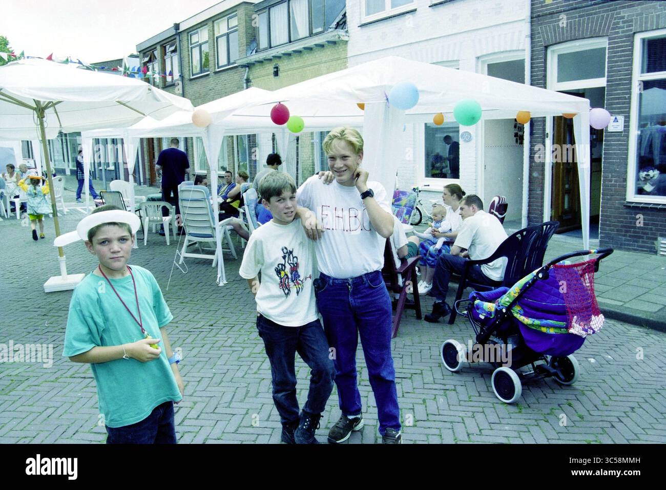 Street party, Javastraat, Haarlem, Javastraat, Paesi Bassi, 02-07-1999 Whizgle News, Dutch Desk, Paesi Bassi, 1950-2000 Una vivace festa di strada che offre ai bambini e agli adulti varie attività sotto tende colorate. Due ragazzi si mettono in posa insieme, uno indossa una maglietta grafica mentre l'altro ha un'espressione giocosa. Nelle vicinanze, altri bambini sono impegnati in giochi, circondati da decorazioni festose e da un passeggino decorato con colori vivaci. Foto Stock