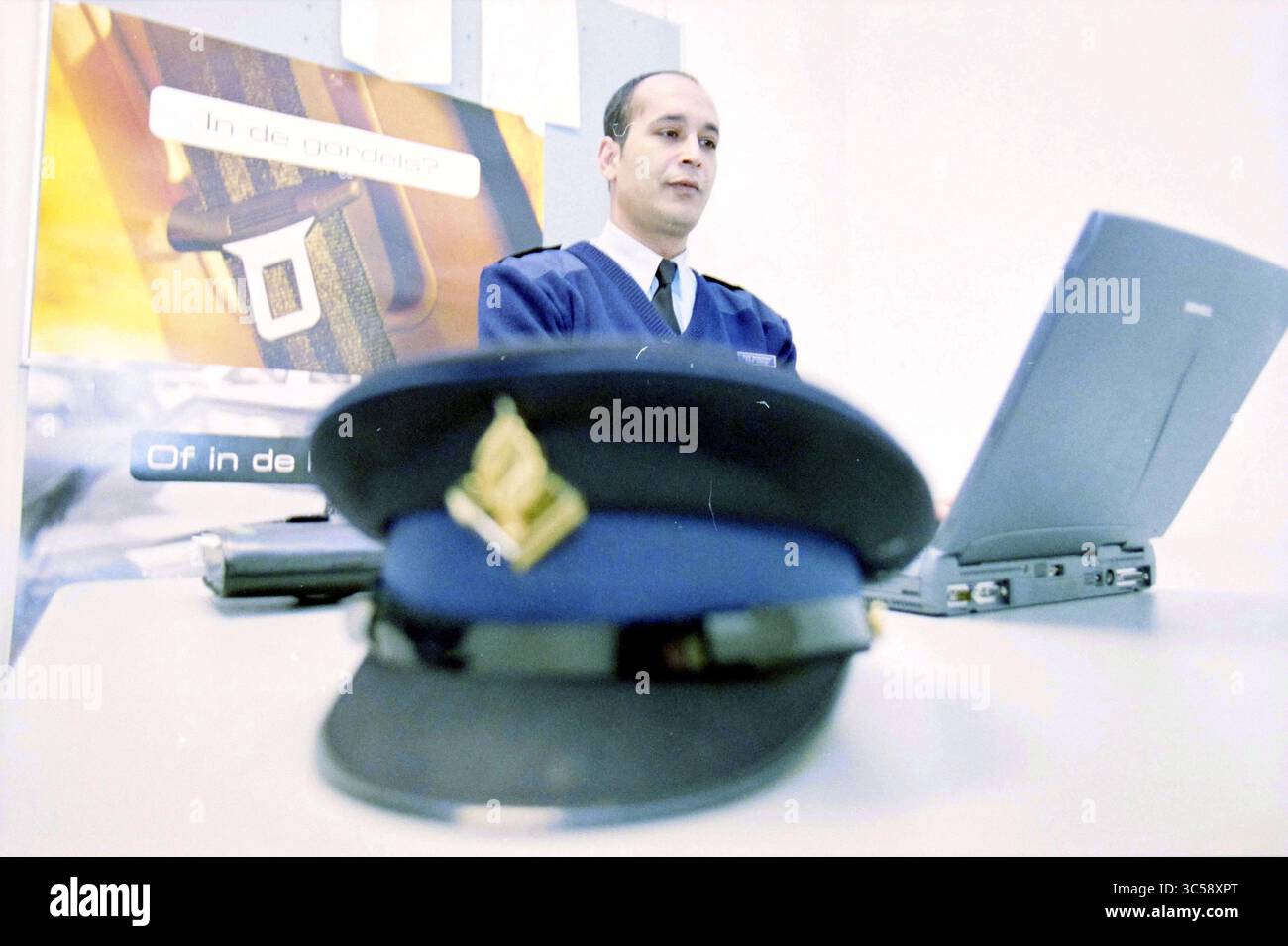 Un poliziotto Whizgle News, Dutch Desk, Paesi Bassi, 1950-2000 Un funzionario in uniforme siede a una scrivania, concentrandosi intensamente su un portatile, mentre un cappellino della polizia è posizionato in primo piano. Lo sfondo presenta materiale promozionale che suggerisce un contesto formale relativo alle forze dell'ordine. Foto Stock