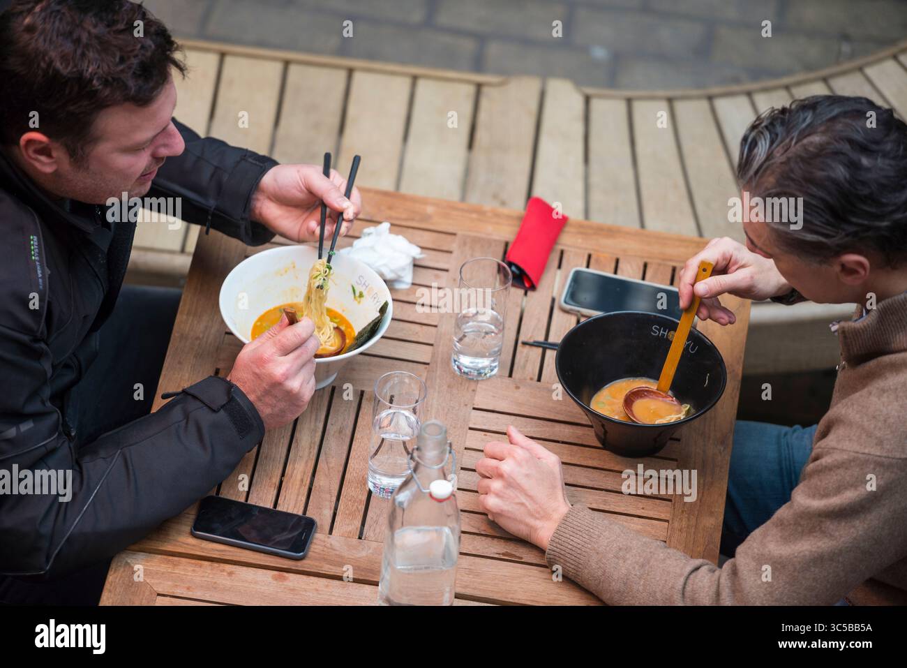 Due uomini che mangiano noodle di ramen all'aperto a un tavolo di legno a Soho, Londra, usando bacchette e cucchiai di zuppa. Foto Stock
