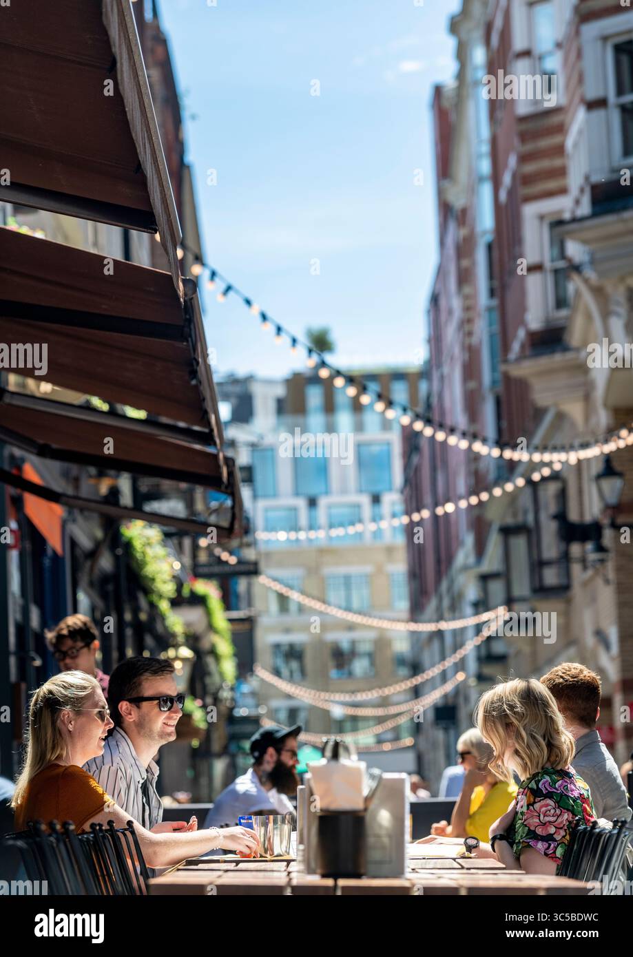 Persone che si godono una cena all'aperto in Kingly Street, Soho, Londra in una giornata estiva soleggiata, con luci sospese decorative che creano un'atmosfera vibrante. Foto Stock