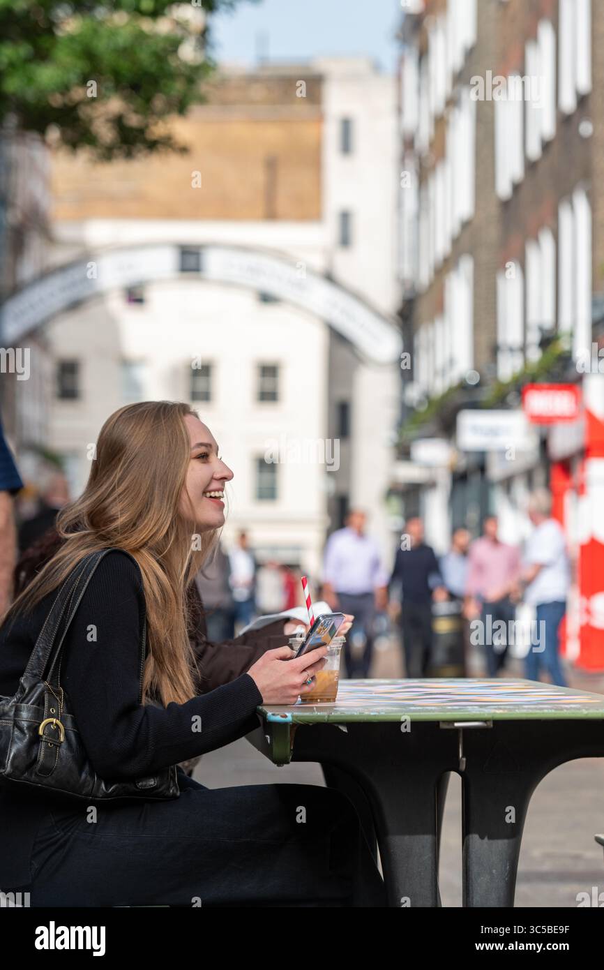 Giovane donna sorridente con un caffè ghiacciato in una caffetteria di strada vicino a Carnaby Street, Soho, Londra in una giornata di sole Foto Stock