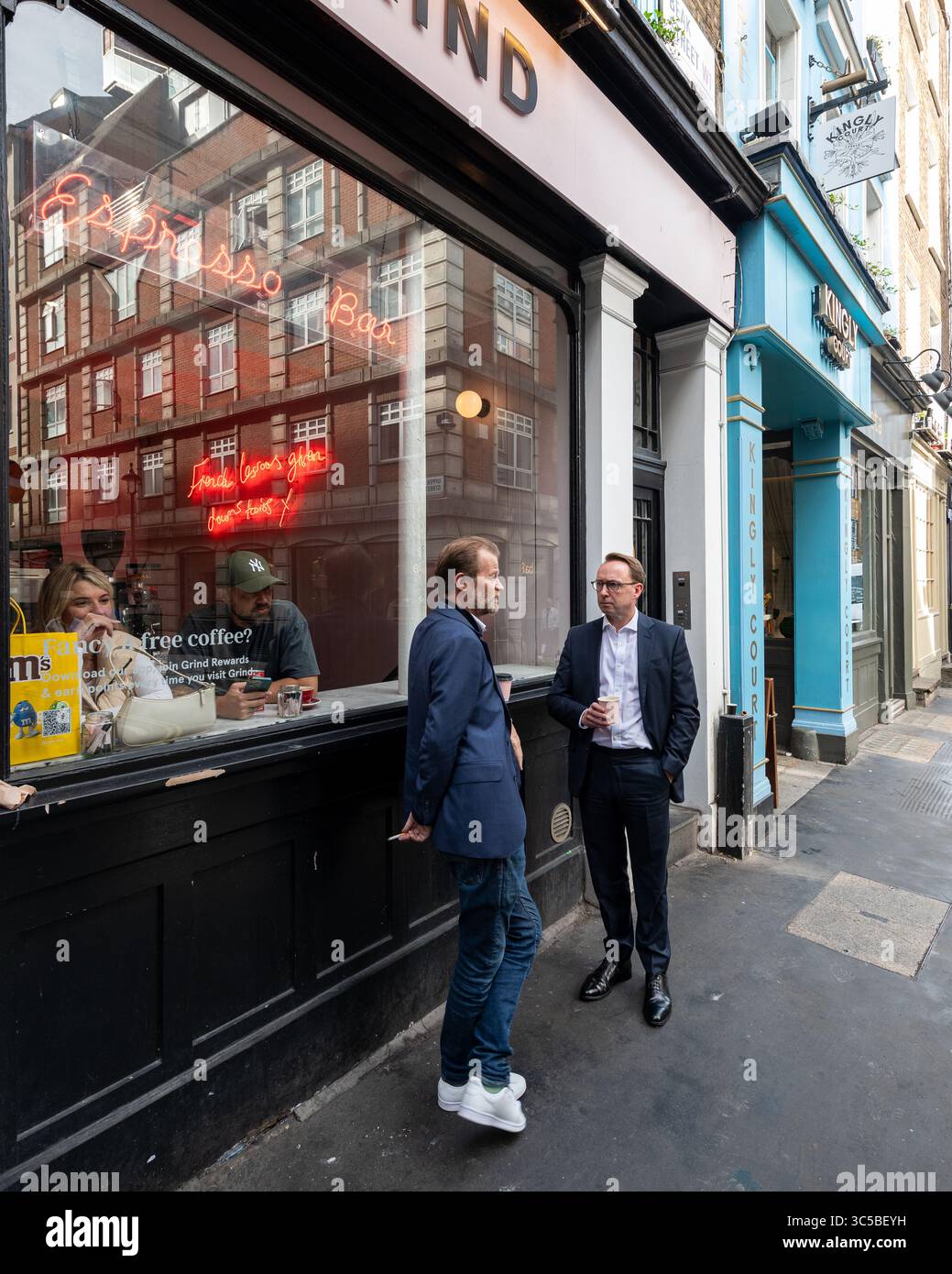 Due uomini parlano fuori dal caffè Grind in Beak Street, Soho, Londra, con riflessi alla finestra e passanti che si godono l'atmosfera della città. Foto Stock