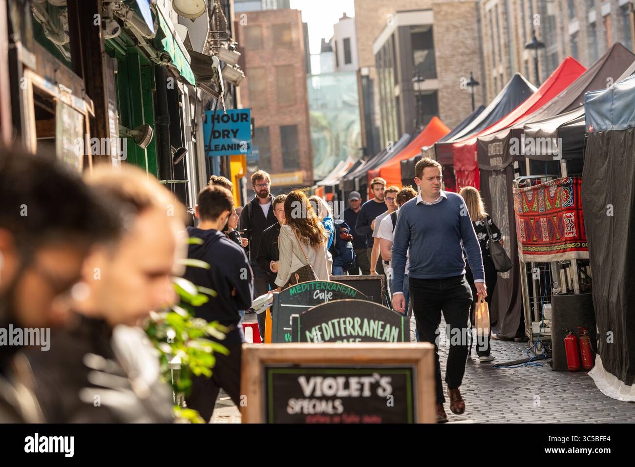 Gli amanti dello shopping e dei pedoni al mercato di Berwick Street a Soho, Londra, in un pomeriggio di sole. Il mercato è noto per le sue vivaci bancarelle e la strada locale Foto Stock