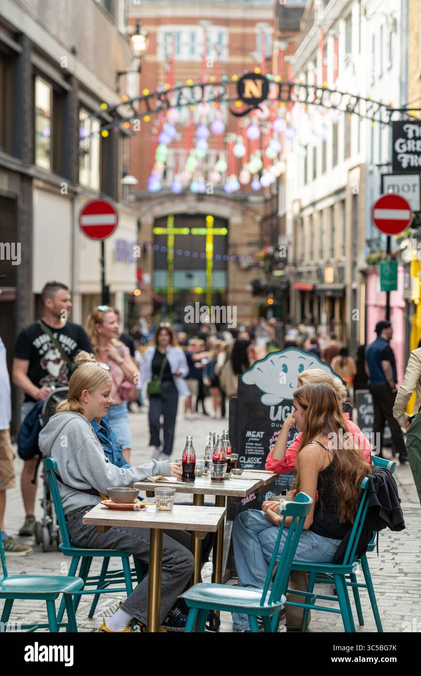 Persone che cenano all'aperto in un ristorante affollato nel quartiere di Newburgh, Soho, con l'iconico cartello ad arco e decorazioni colorate di strada alle spalle. Foto Stock