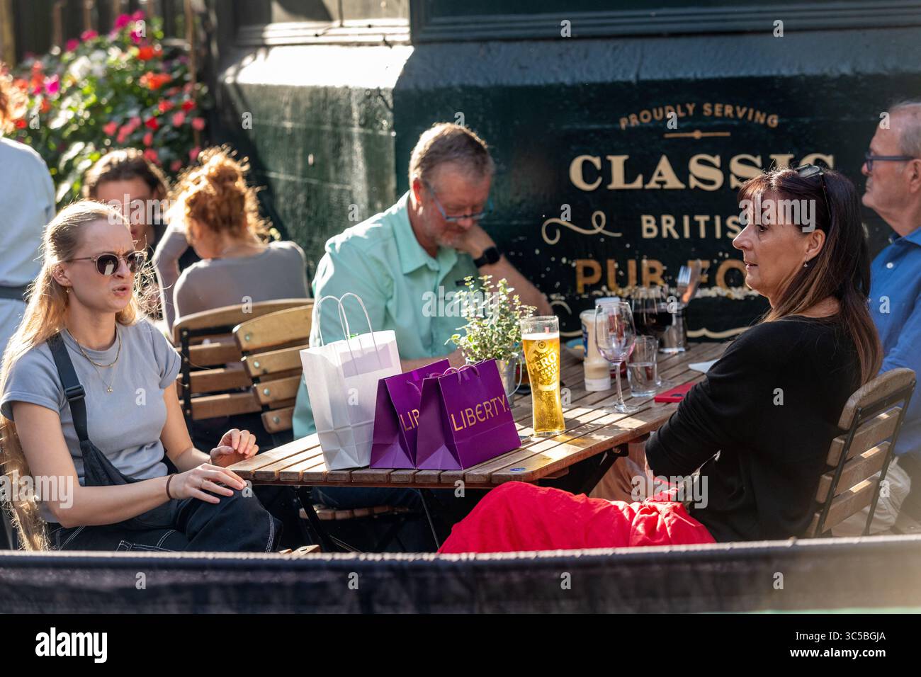 Persone che si godono un pomeriggio di sole fuori da un classico pub britannico a Soho, Londra, con borse dello shopping Liberty e bevande sul tavolo Foto Stock
