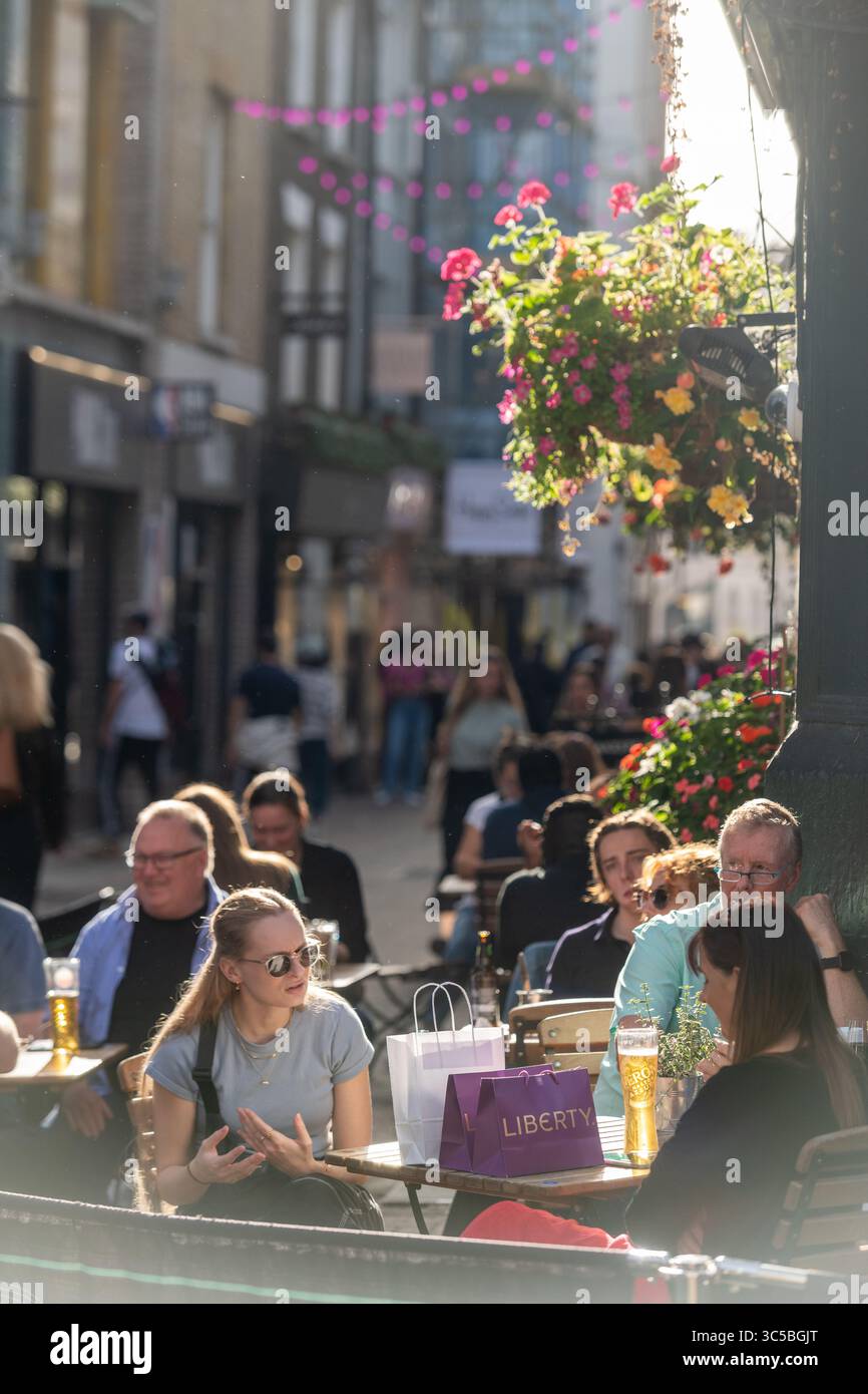 Persone che amano bere e fare shopping all'aperto in un pomeriggio di sole a Soho, Londra, con borse Liberty e vivaci esposizioni floreali Foto Stock