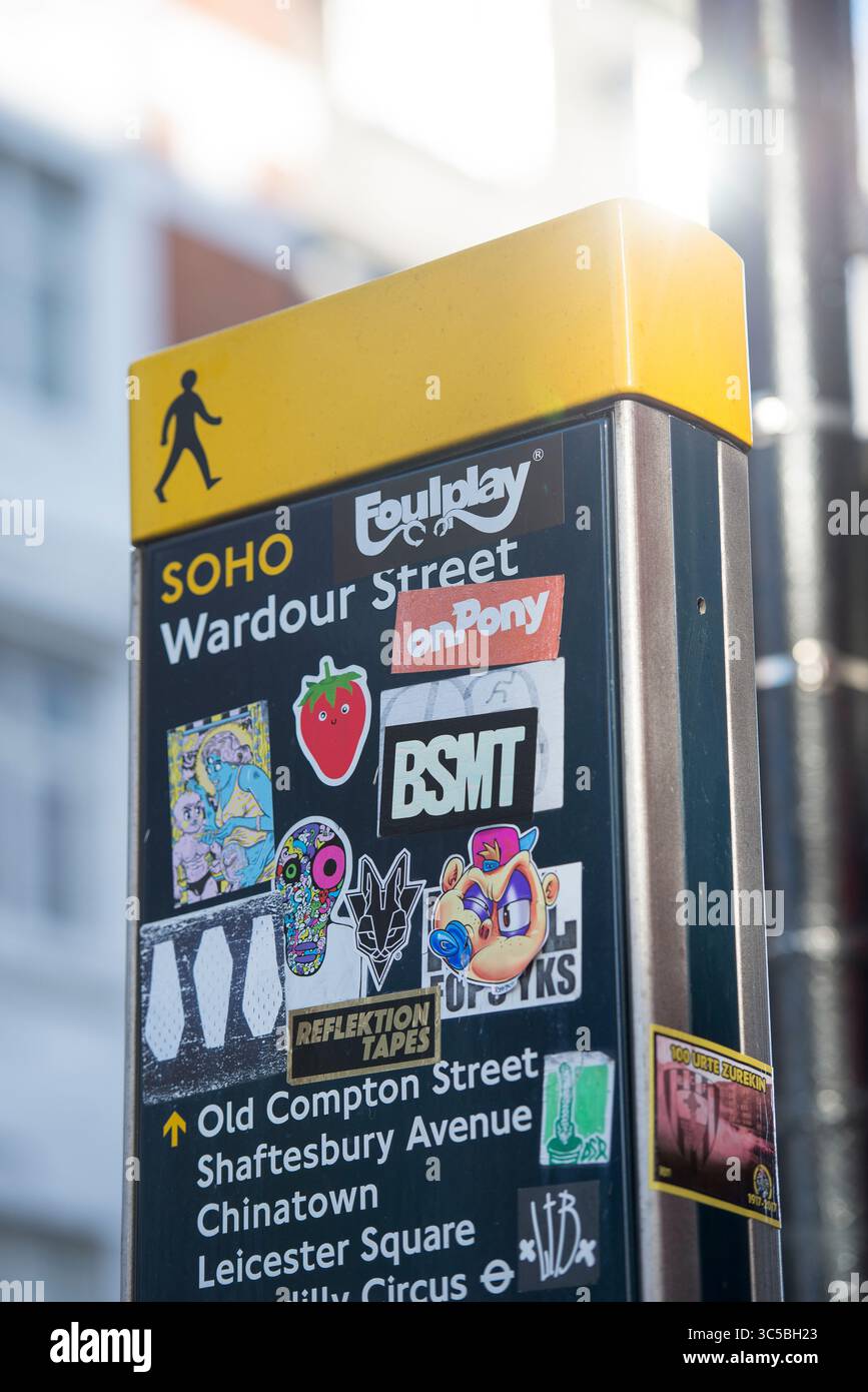 Cartello pedonale direzionale per Wardour Street a Soho, Londra, coperto da adesivi colorati, con edifici della città e luce del sole sullo sfondo. Foto Stock