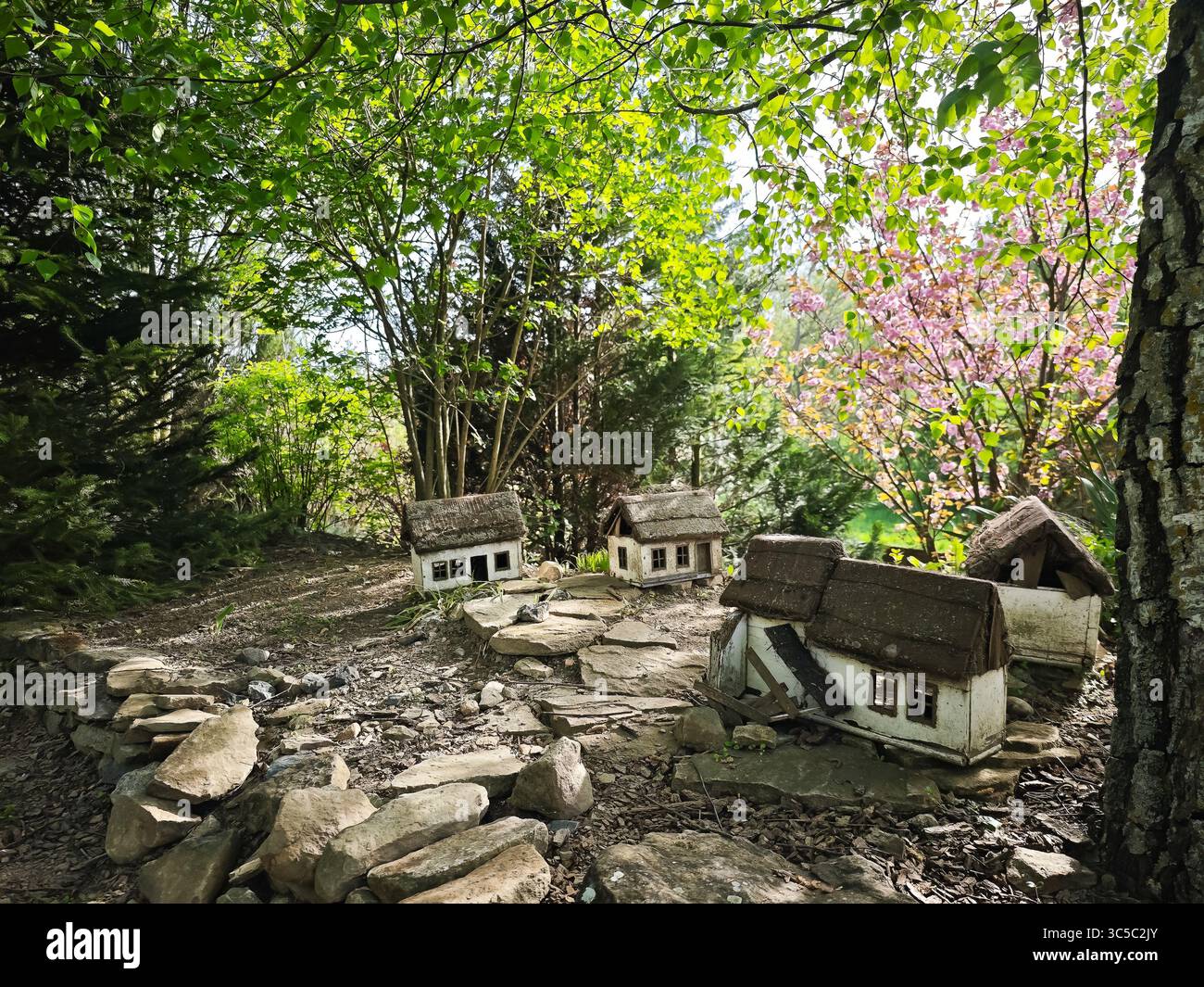 Cottage rustici in miniatura con tetti di paglia e piastrelle collocati in un giardino forestale, circondati da vegetazione primaverile e fiori rosa fiorenti. Foto Stock