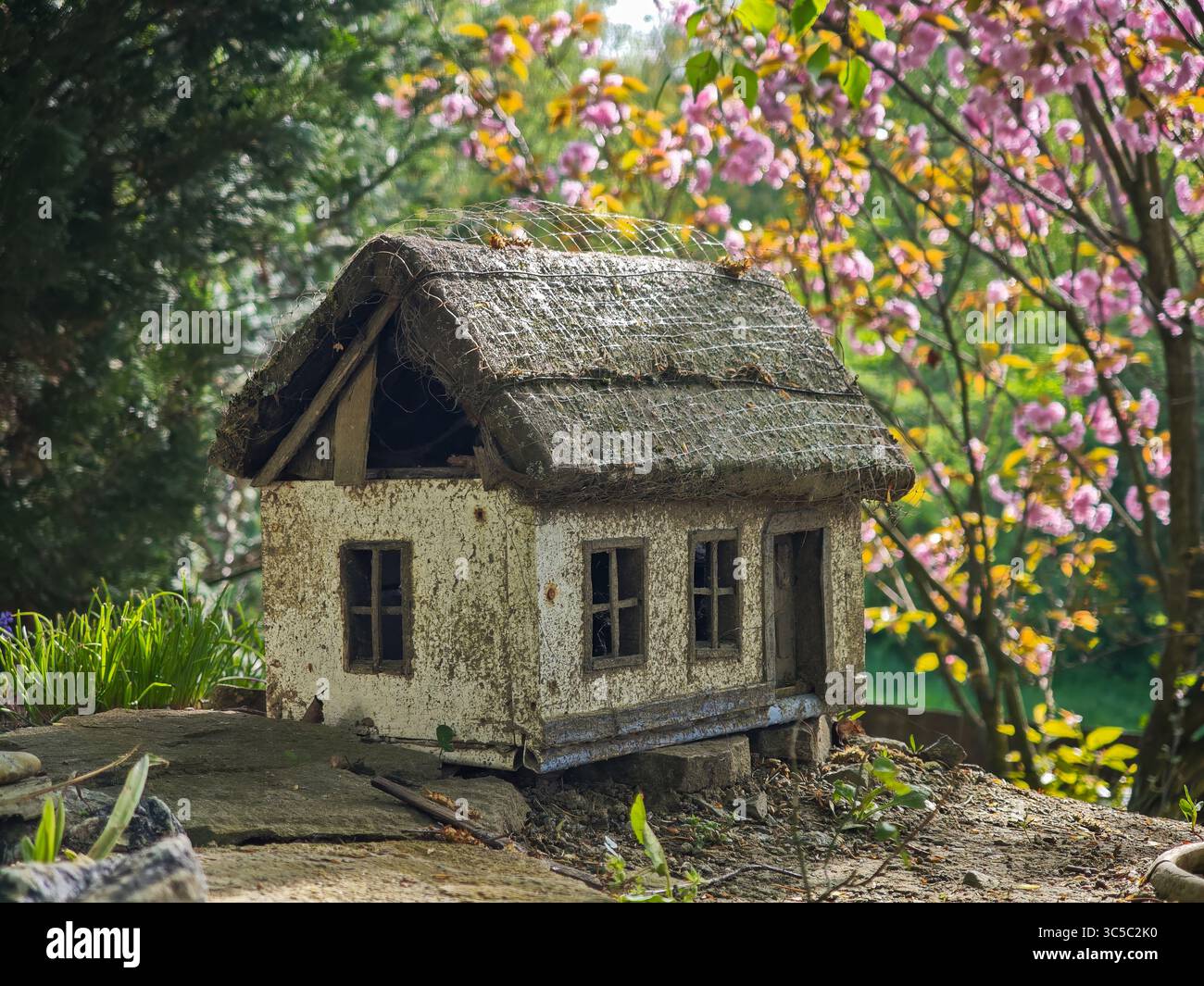 Primo piano di una casa in miniatura rustica con tetto in paglia, circondata da fiori primaverili e vegetazione verde in un giardino. Foto Stock