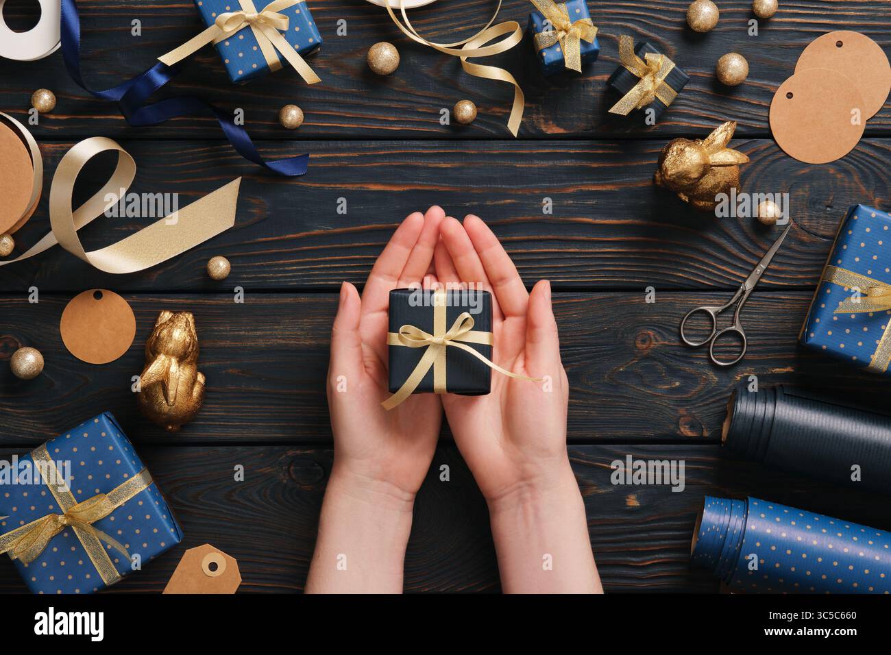 Le mani reggono una scatola regalo blu con nastro d'oro circondata da articoli da regalo su uno sfondo blu in legno Foto Stock