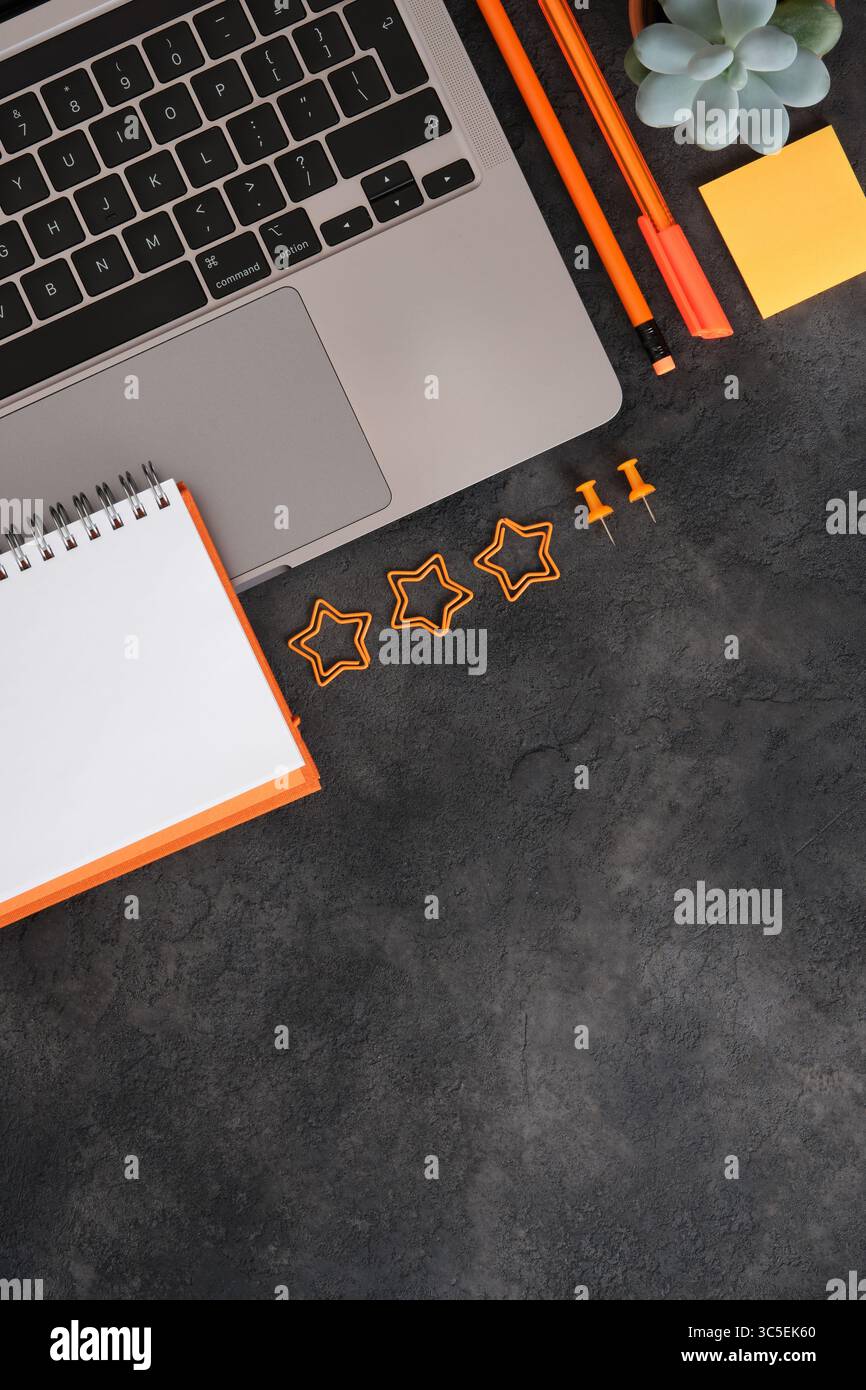 Spazio di lavoro con laptop, cancelleria arancione, blocco note e succulento su sfondo scuro. Copia spazio. Foto Stock