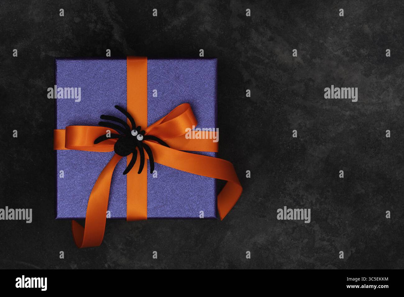 Regalo di Halloween con carta da imballaggio viola, nastri arancioni, ragno nero su sfondo scuro. Copyspace. Foto Stock