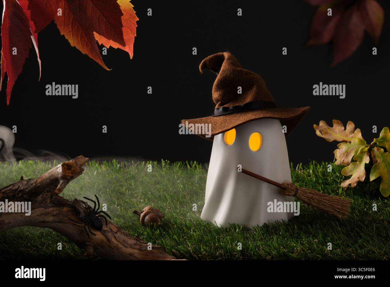 Simpatico fantasma di Halloween con cappello da strega e scopa, occhi accesi sull'erba verde con foglie autunnali e ragno, spaventosa scena autunnale Foto Stock