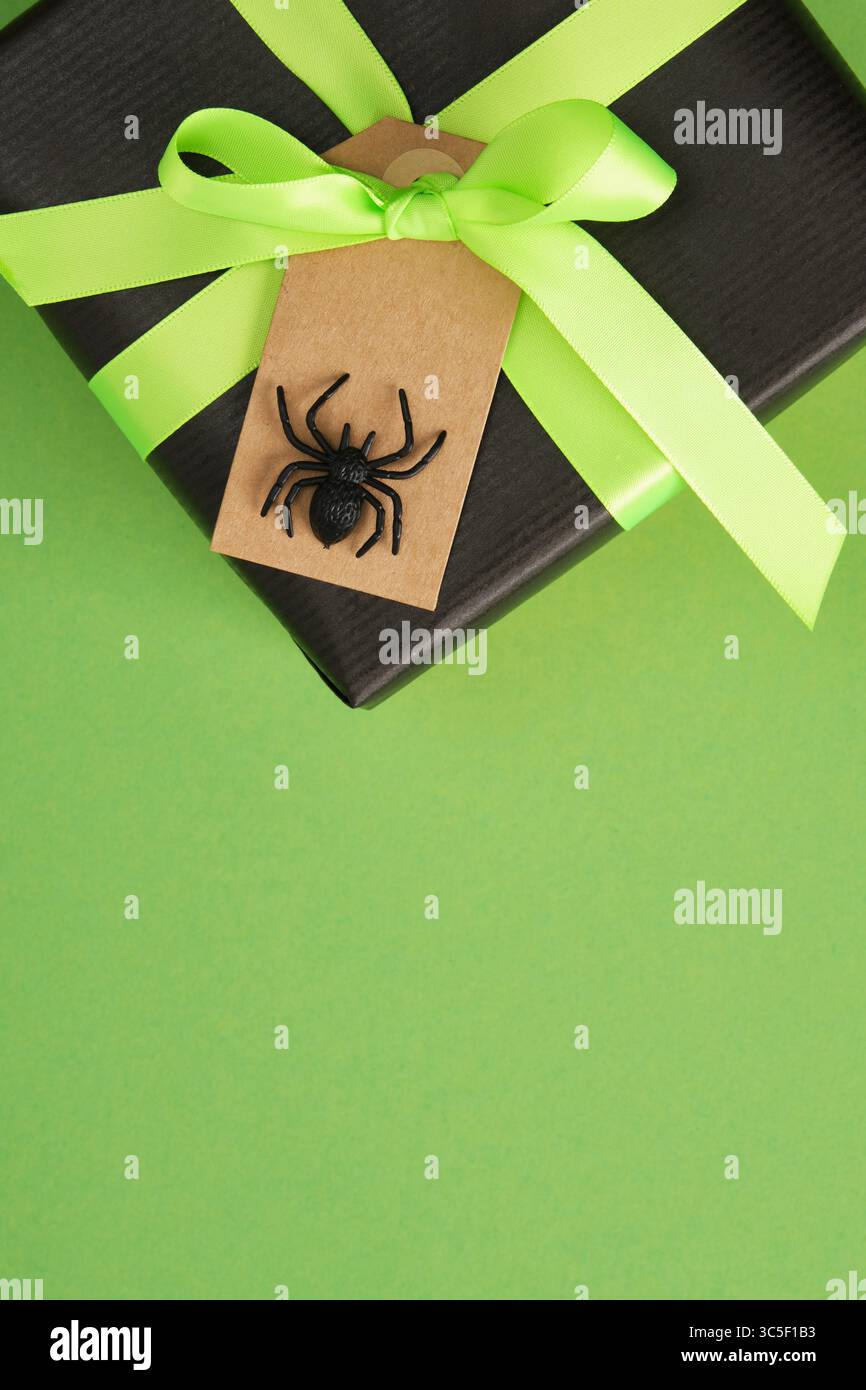Regalo di Halloween con ragno nero, nastro verde neon e carta da imballaggio nera su sfondo verde. Banner verticale, spazio per il testo. Foto Stock