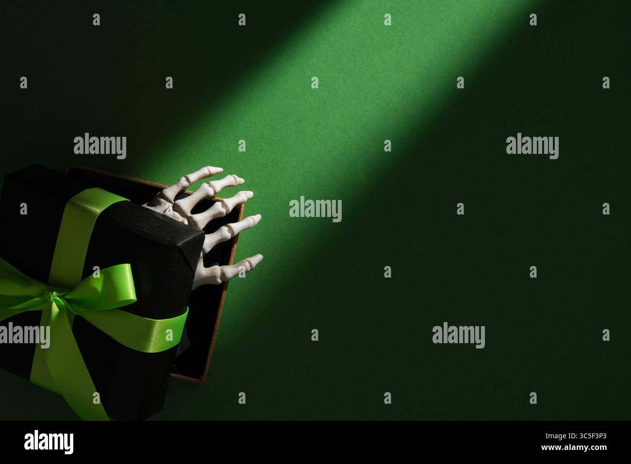 Mano di scheletro che emerge dalla confezione regalo nera con nastro verde su sfondo verde scuro, misterioso concetto di regalo di Halloween. Copyspace. Foto Stock