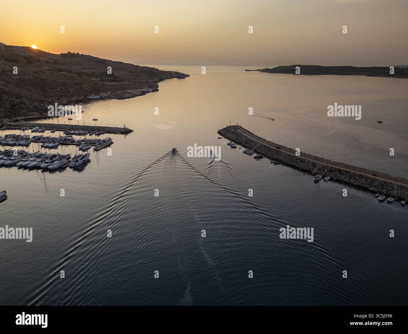 Vista aerea di un tranquillo ingresso al porto dove il sole radioso bacia l'orizzonte, proiettando un bagliore dorato sulle barche, Gajnsielem, Malta. Foto Stock