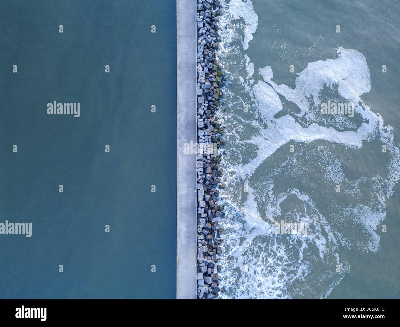 Veduta aerea di un molo di cemento che divide il tranquillo e profondo mare blu dalle onde spumeggianti e turbolente che si infrangono contro il frangiflutti di pietra, Wijk aan Zee, Olanda settentrionale, Paesi Bassi. Foto Stock