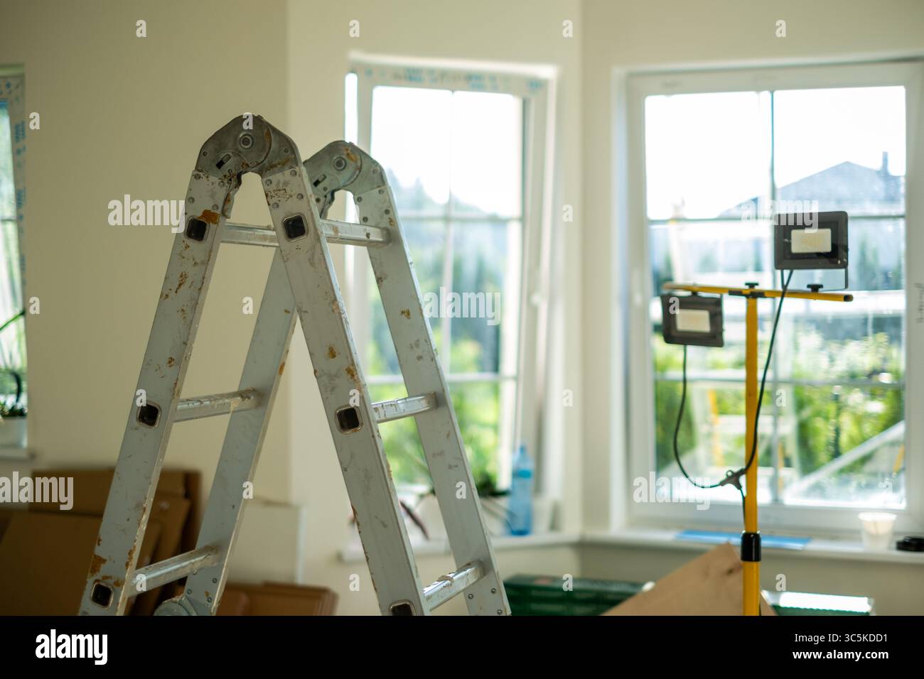 La scala in metallo si trova in una sala ben illuminata con grandi finestre, circondata da strumenti e materiali, creando un'atmosfera invitante per lavori di ristrutturazione della casa Foto Stock