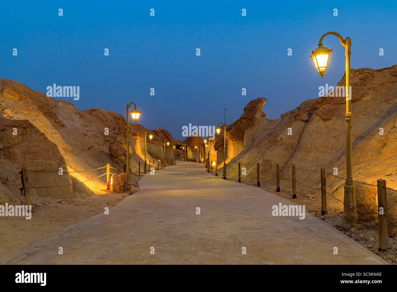 Strada illuminata e arco che conducono agli edifici tradizionali delle grotte rocciose di montagna di Jabal al Qarah di notte nell'oasi di al Ahsa, Arabia Saudita Foto Stock