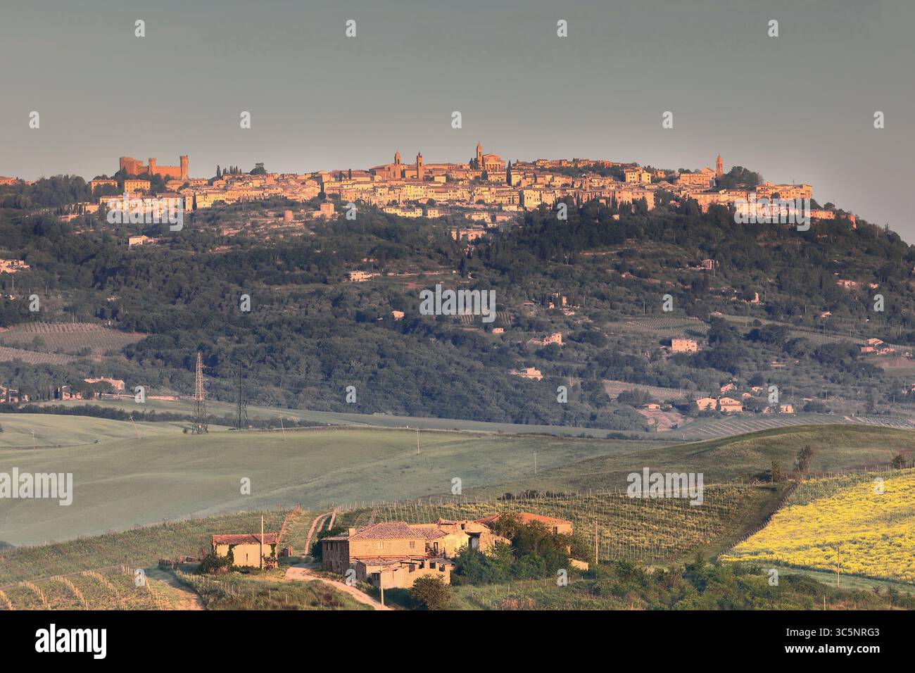 152 calda luce dell'alba sopra Montalcino, su una collina con terreni agricoli e boschi ai suoi piedi, vista verso ovest dalla SS2-via Cassia. Toscana-Italia. Foto Stock