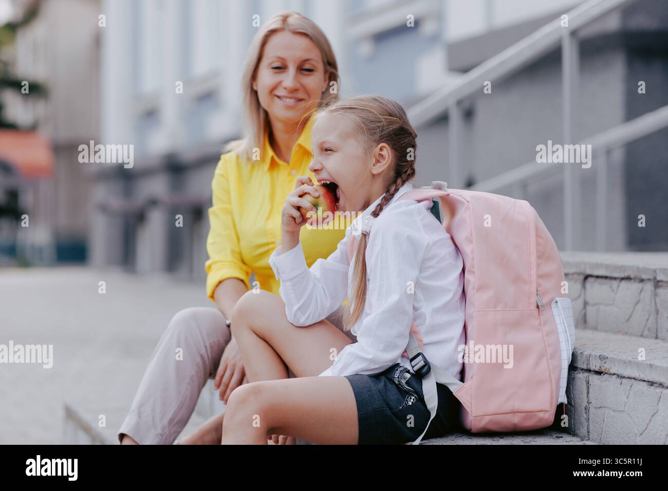 Il giovane studente mangia la mela con lo zaino e la madre nelle vicinanze prima della lezione, mostrando abitudini sane al mattino di ritorno a scuola. Foto Stock