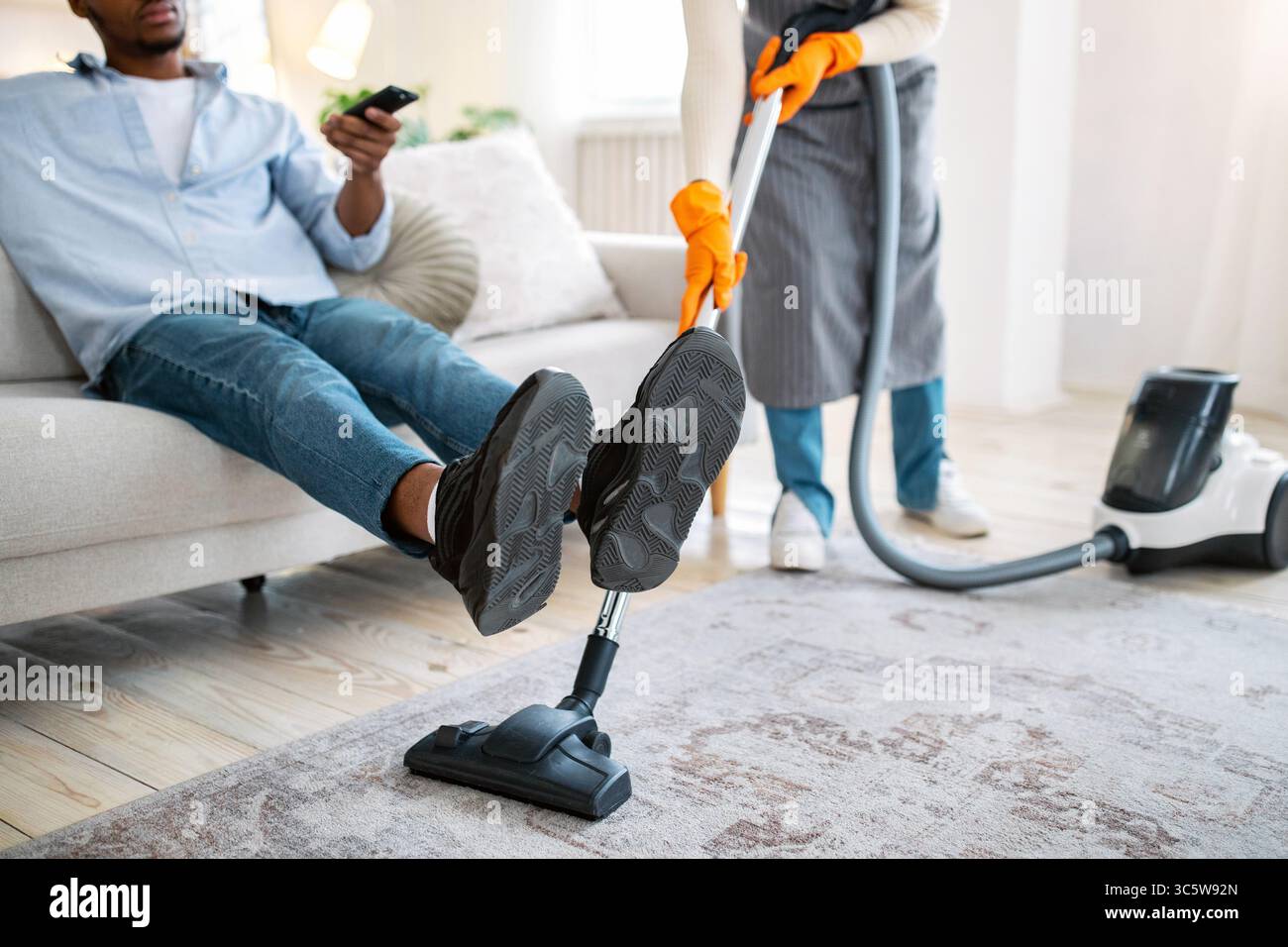 Signora nera che aspira il pavimento mentre suo marito guarda la TV, non aiutandola con i doveri della famiglia, vista ritagliata Foto Stock
