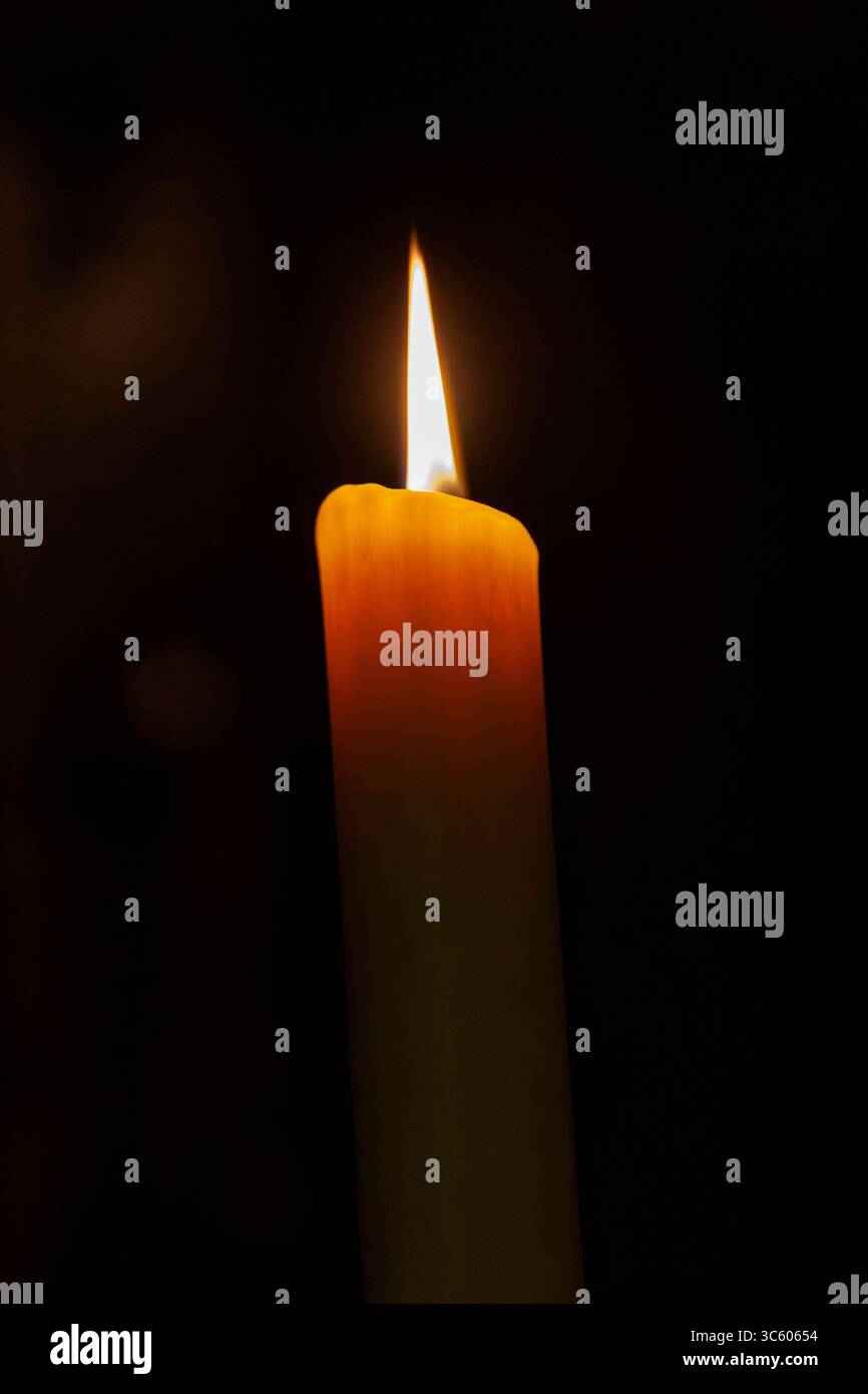 Una singola candela è alta e la sua fiamma tremola con la luce calda che emette una luce soffusa Foto Stock
