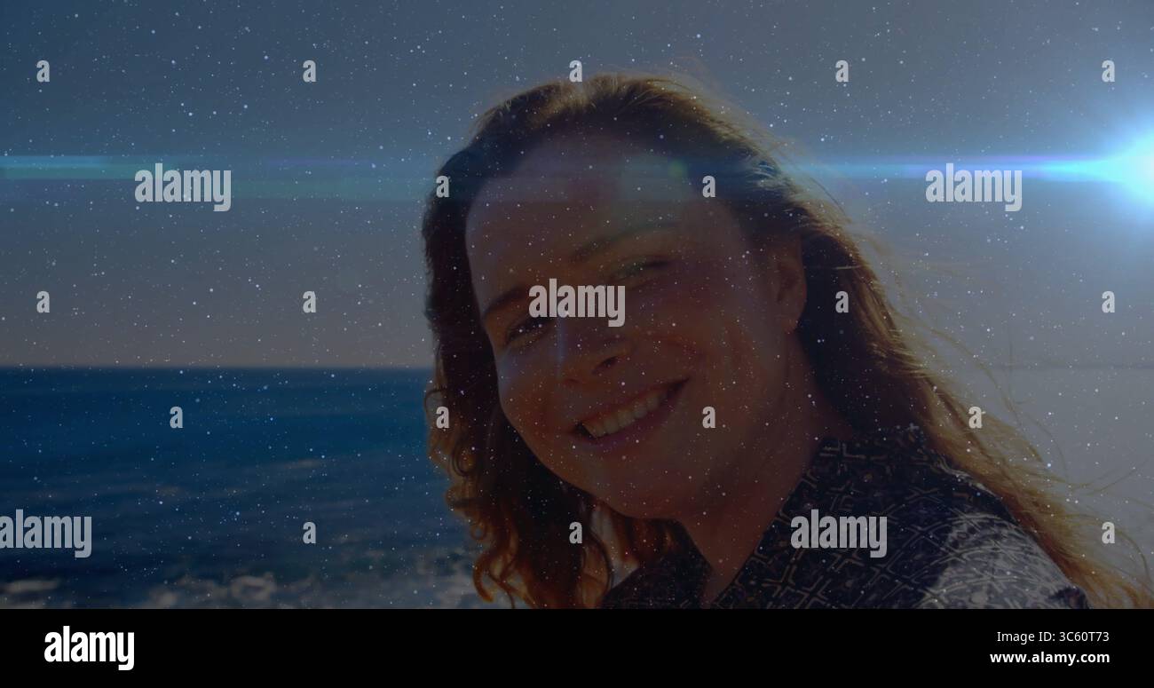 Donna sorridente che indossa una camicia scura a motivi geometrici che guarda la costa dell'oceano sotto un bagliore luminoso Foto Stock
