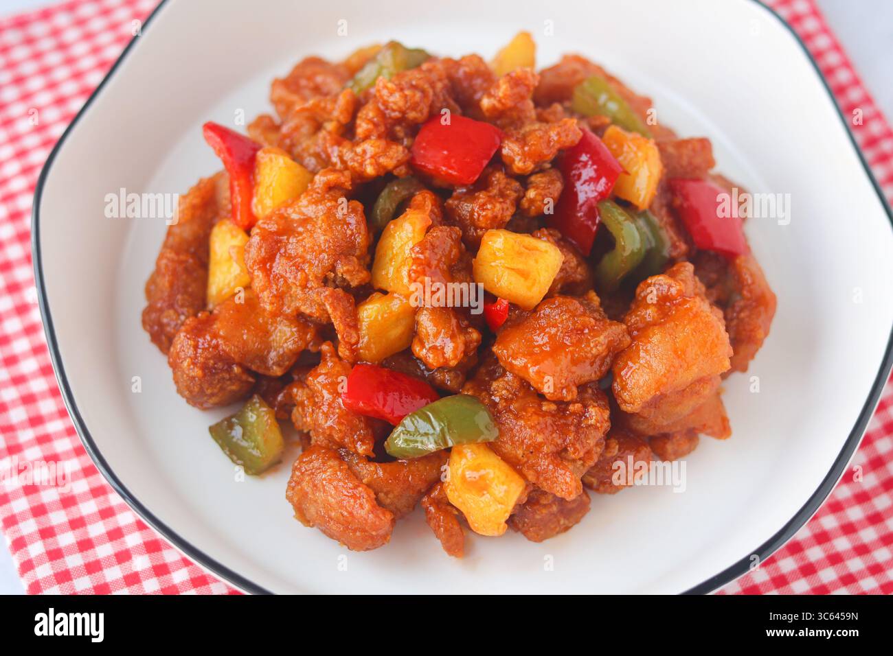 Primo piano di pollo agrodolce fatto in casa con peperoni e pezzi di ananas, servito su un piatto bianco con un tovagliolo a scacchi rossi. Foto Stock