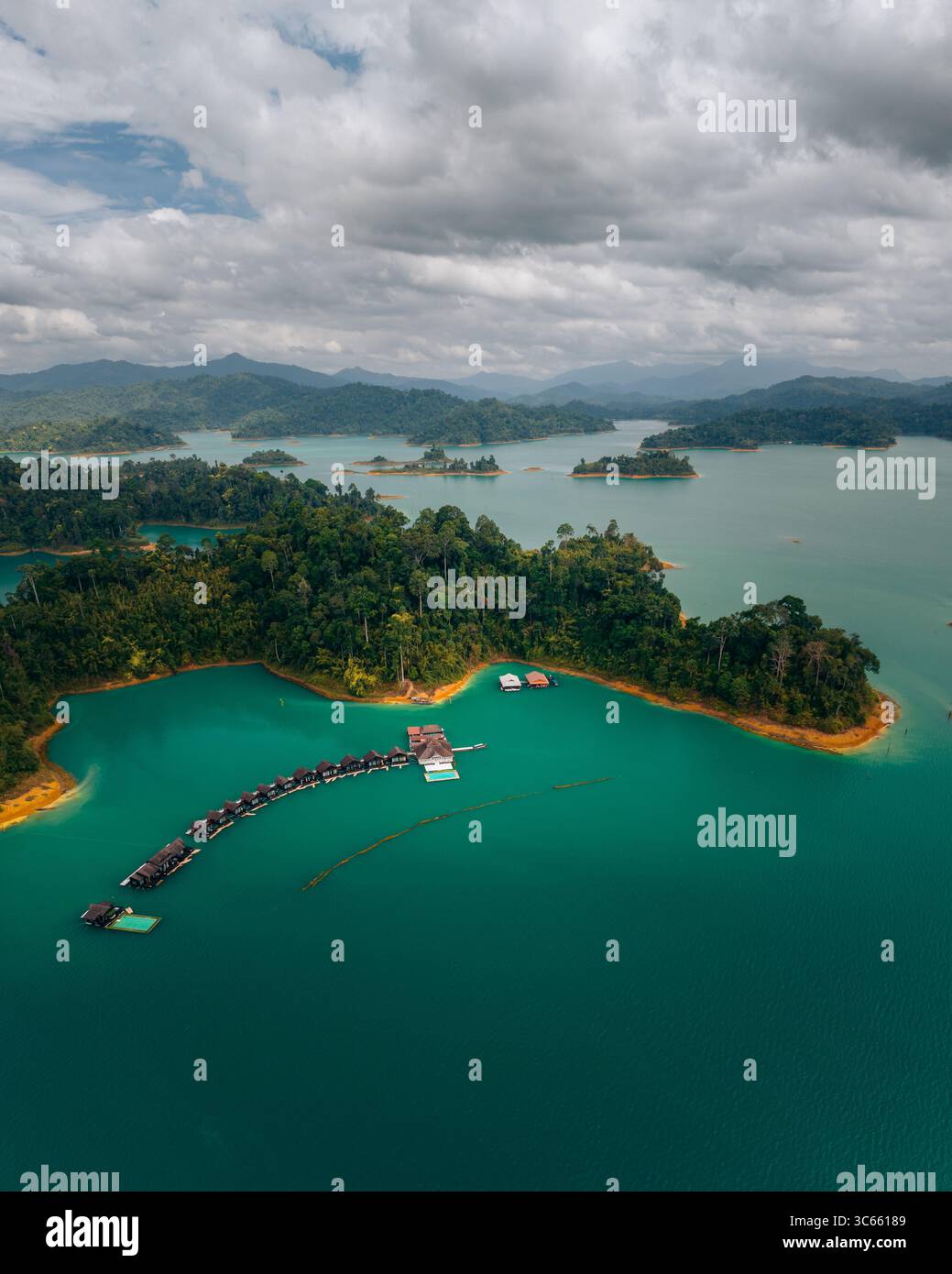 Vista aerea di un vivace lago turchese punteggiato da isole verdeggianti, collegate da bungalow galleggianti, sotto un cielo di nuvole vorticose, Parco Nazionale di Khao Sok, Surat Thani, Thailandia. Foto Stock