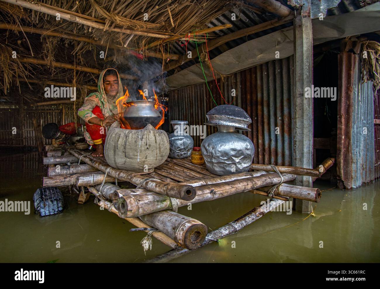Bogura, Bangladesh - 15 luglio 2019: Vista di una donna che cucina su un fuoco aperto in mezzo alle inondazioni all'interno di un'umile abitazione, dove il metallo ondulato e il tetto di paglia incontrano le profondità torbide. Foto Stock
