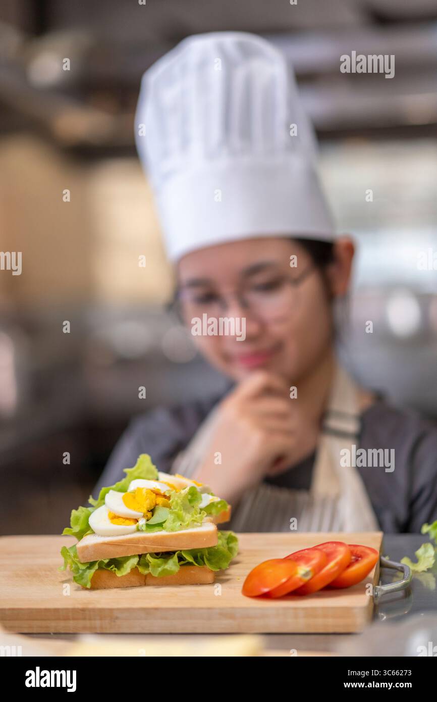 Una giovane chef donna in uniforme esamina attentamente un panino che ha preparato, mostrando attenzione, creatività e abilità culinarie in una cucina commerciale Foto Stock
