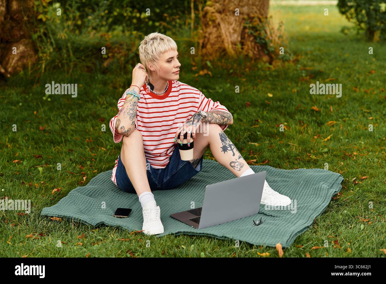 Una giovane donna con i tatuaggi si rilassa su una coperta, sorseggiando un caffè e lavorando al suo laptop all'aperto. Foto Stock