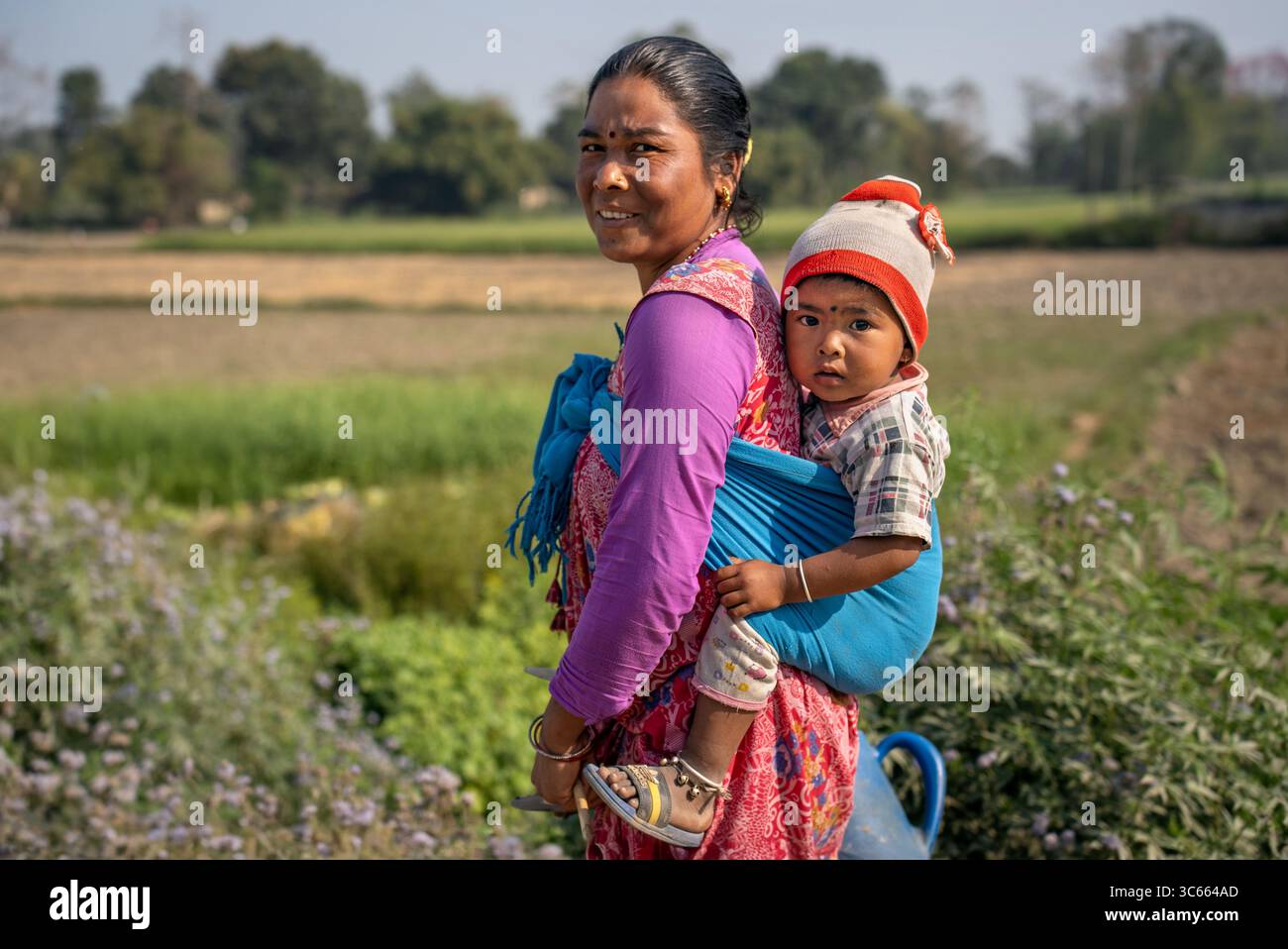 Dhangadhi, Nepal - 03 marzo 2023: Vista di una madre in un vivace sari a fantasia, che trasporta il suo bambino in una fionda blu, nel mezzo della lussureggiante vegetazione del co Foto Stock