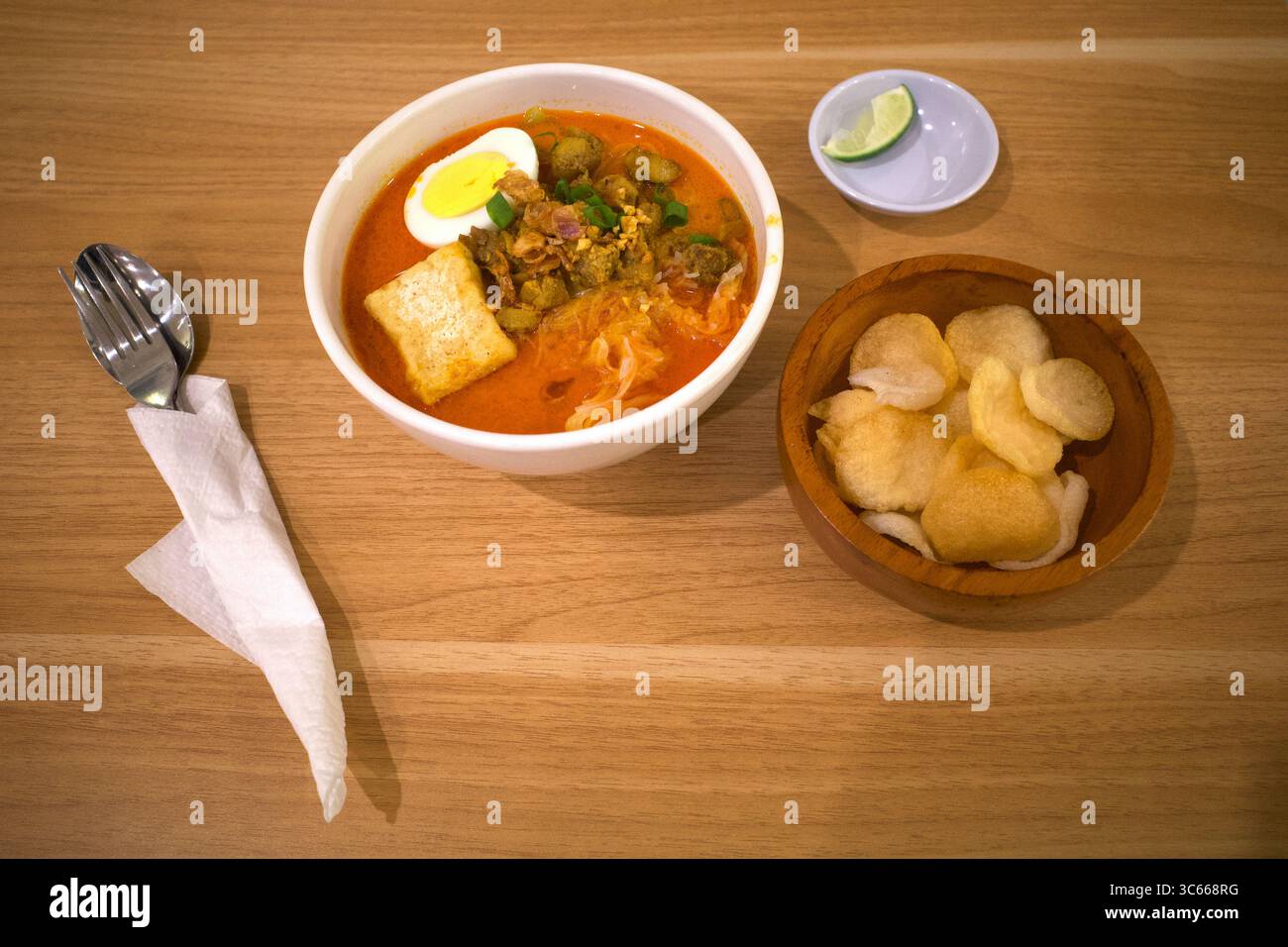 Una ciotola di tagliatelle al curry Laksa con uovo, una fetta di lime e cracker, cucina tradizionale del Sud Est Asiatico su un tavolo di legno. Foto Stock
