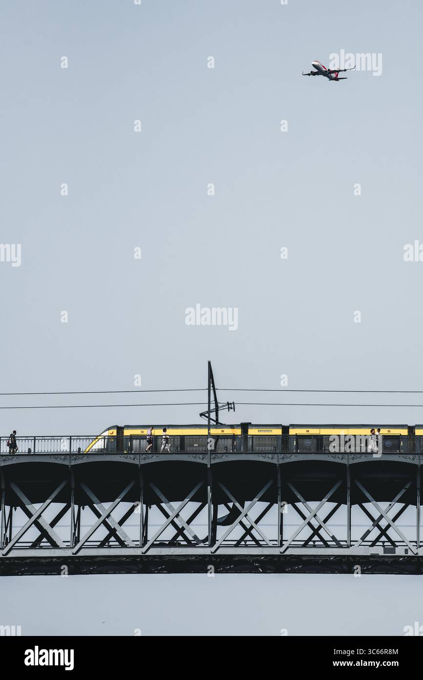 Porto, Portogallo - 17 giugno 2022: Veduta di un moderno treno giallo che attraversa il ponte metallico Dom Luís i mentre un aereo vola sopra, una miscela di modi di trasporto contro un cielo tenue. Foto Stock