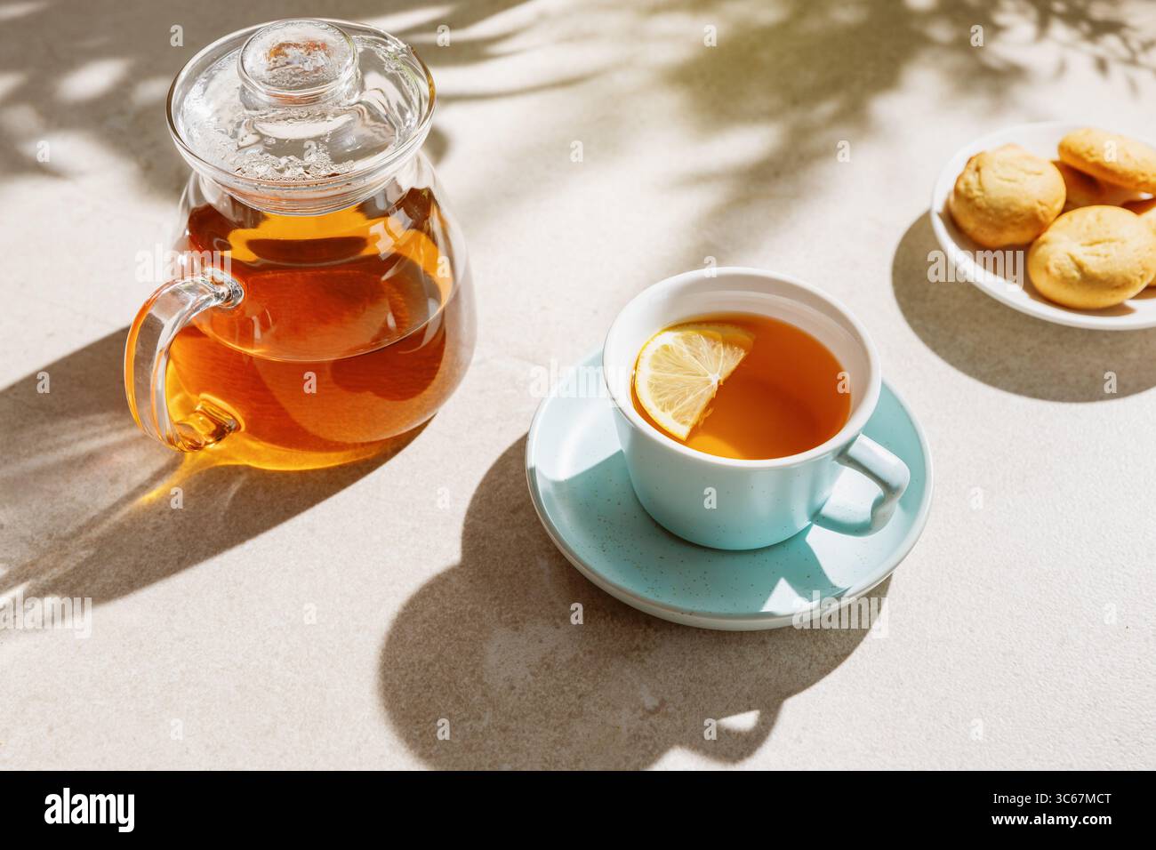 Tazza di tè fresco con una fetta di limone e teiera su un tavolo illuminato dal sole. Foto Stock