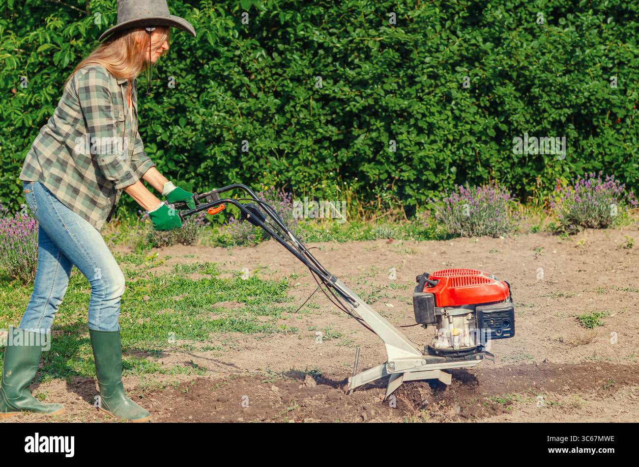 Contadina in cappello e jeans ara il terreno con coltivatore di benzina manuale in giardino, preparando terreni per la semina. Concetto di agricoltura. Foto Stock