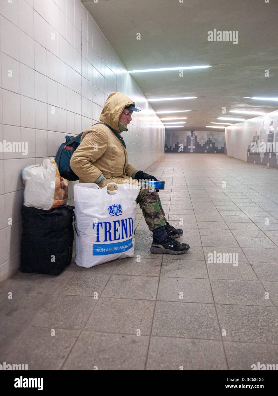 Una persona siede da sola su una panchina in un tunnel pedonale sotterraneo con piastrelle bianche, evidenziando le questioni sociali urbane e l'uso degli spazi pubblici. Foto Stock
