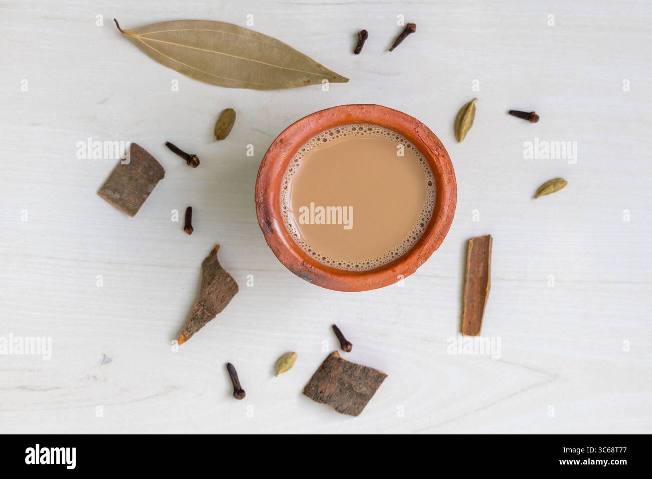 Vista dall'alto di una tazza di masala chai, o Kadak Chai indiano, servita in una tazza di argilla su una superficie di legno, circondata da spezie aromatiche come cardamomo, chiodi di garofano, Foto Stock