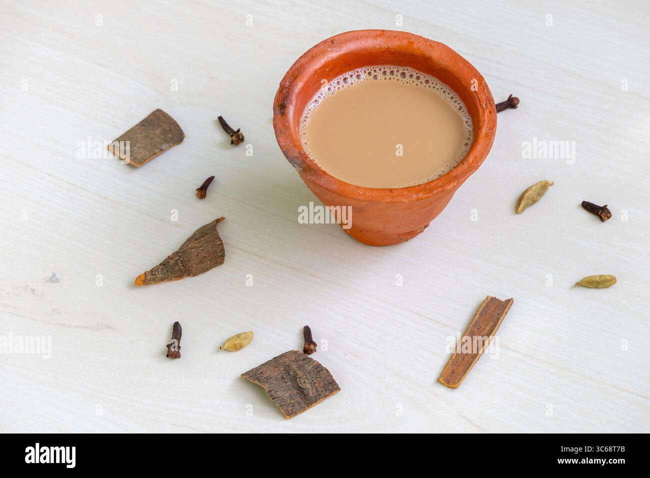 Una tazza di masala chai appena preparato, o Kadak Chai indiano, versata in una tradizionale tazza di argilla, appoggiata su una superficie di legno. Sparsi intorno ci sono le A. Foto Stock
