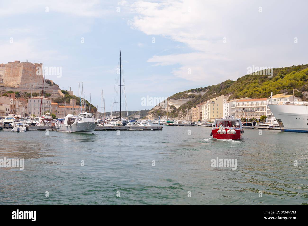 Tranquillo paesaggio marittimo che mostra un porto vivace con barche attraccate vicino a edifici affascinanti, circondate da lussureggianti colline verdi e un cielo sereno. Foto Stock