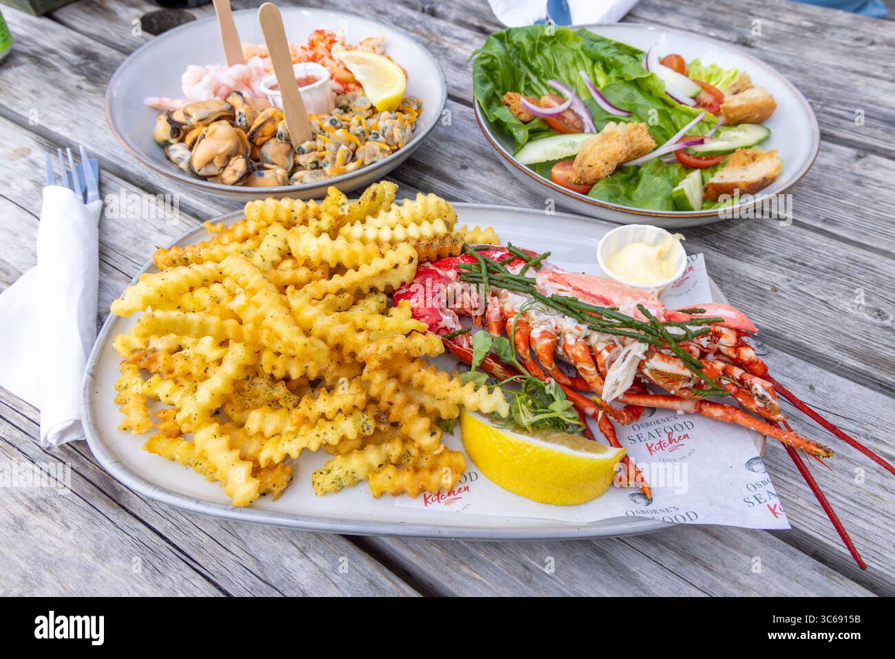 Pranzo presso il ristorante di pesce Osbournes a Leigh-on-Sea, Essex, Regno Unito. Un tavolo estivo con aragosta, patatine fritte tagliate a rughe e piatto di pesce Foto Stock