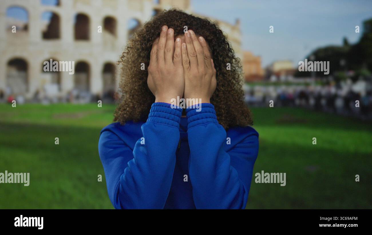 Donna rivela viso e capelli ricci con le mani in felpa blu di fronte al colosseo romano; esplorazione scoperta curiosità. Foto Stock