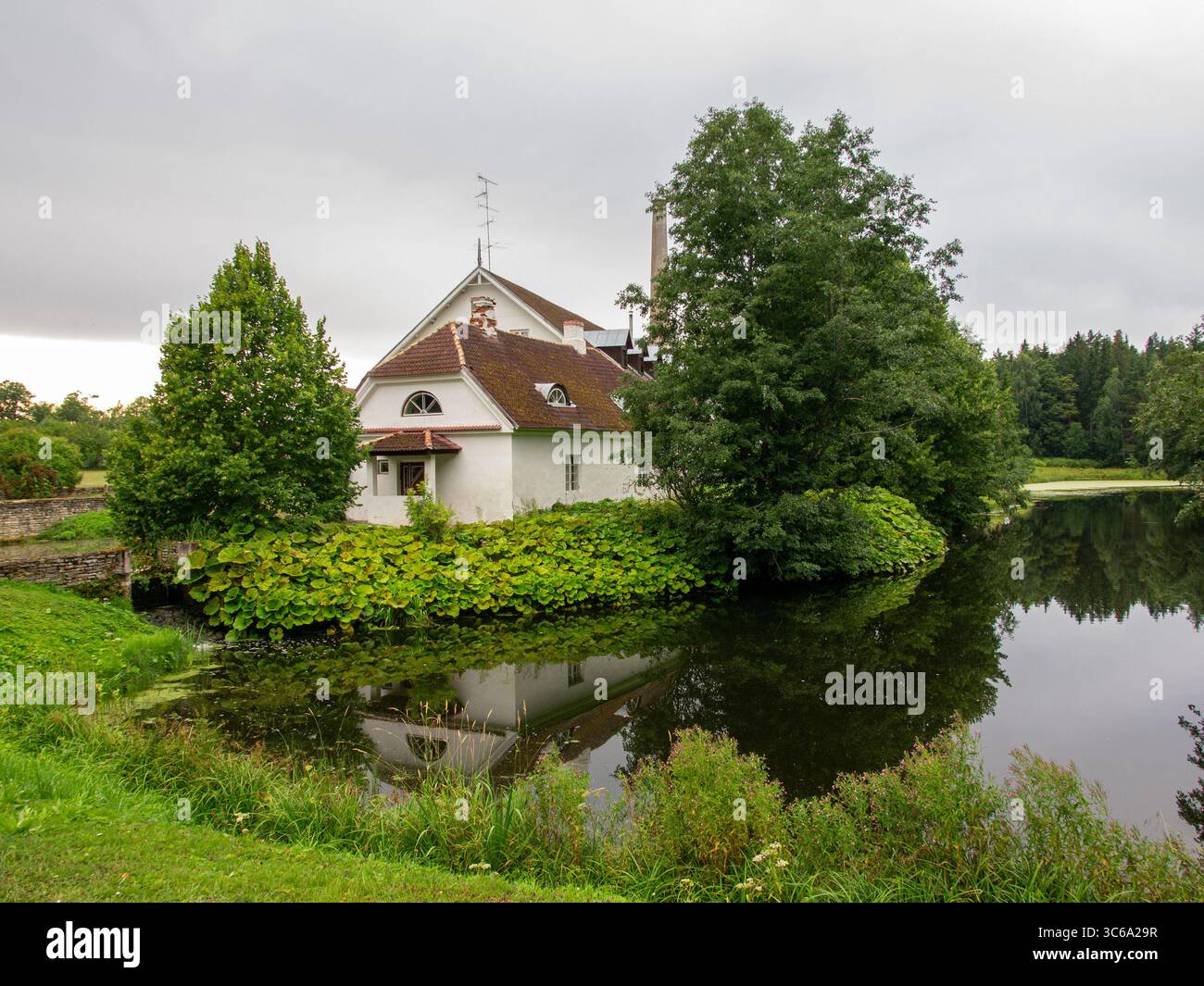 Un edificio storico nella tenuta di Palmse Manor con pareti bianche e tetto rosso situato accanto a uno stagno, circondato da lussureggianti alberi verdi Foto Stock