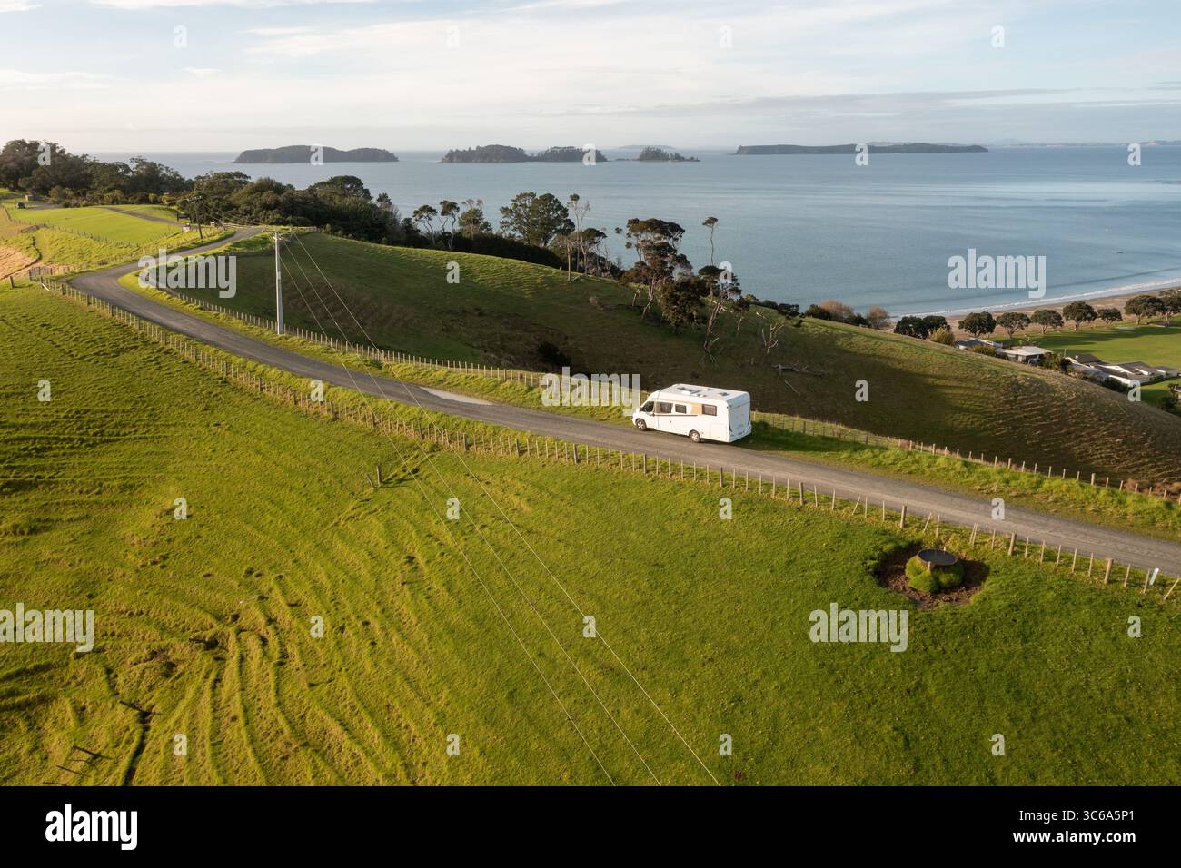 La vista aerea di un camper bianco luminoso si snoda lungo una strada costiera vicino a una vibrante collina verde che si affaccia sul tranquillo oceano blu, Whangarei, Northland Region, nuova Zelanda. Foto Stock