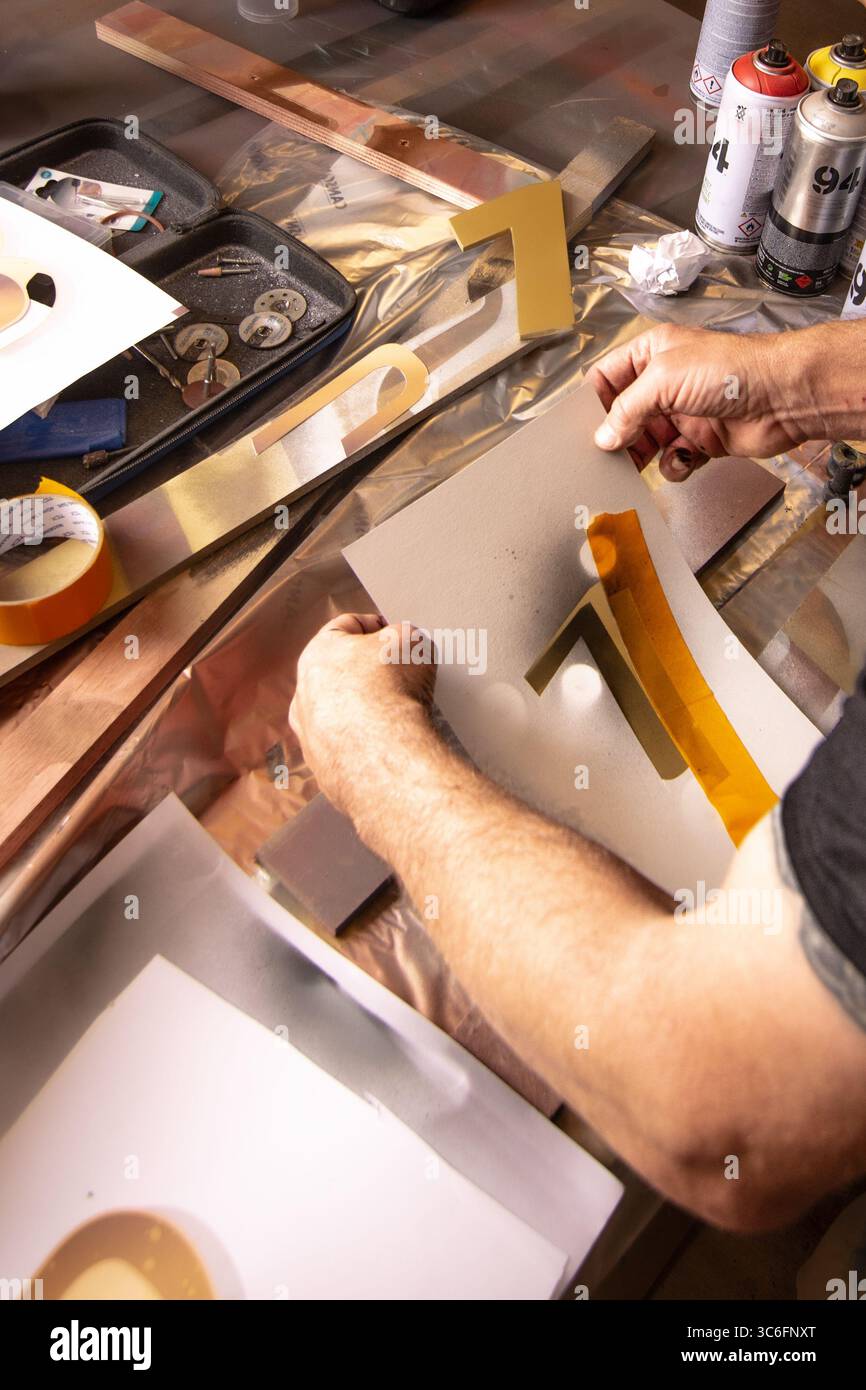 Primo piano di un artista che realizza stencil a numeri d'oro con vernice spray su un ampio tavolo da officina. Ideale per i temi della creatività, del fai da te e dell'arte pratica. Foto Stock