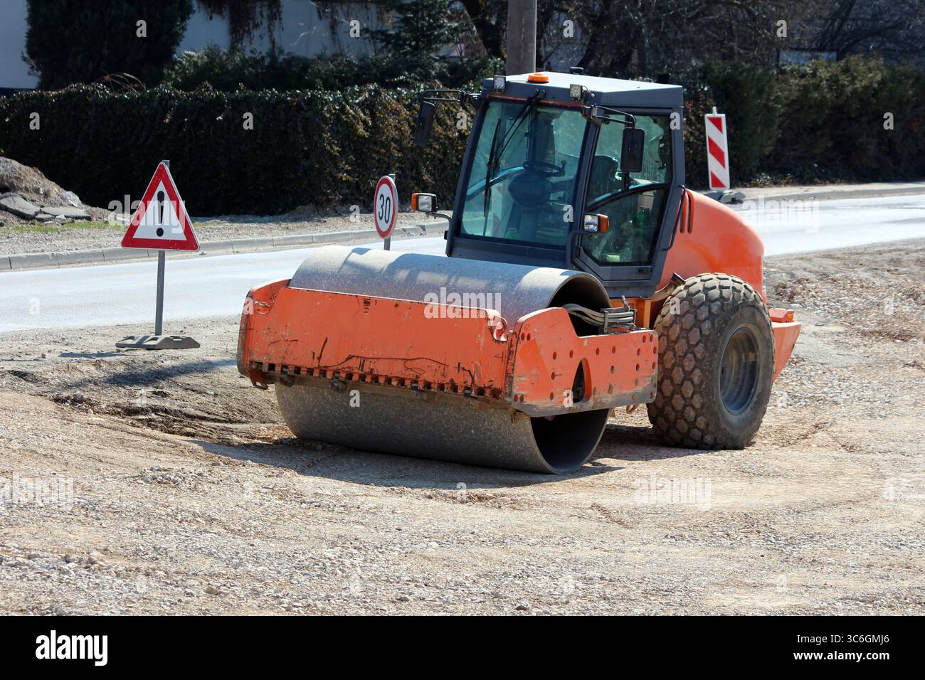 Un rullo vibrante arancione a tamburo singolo compatte ghiaia e terreno in un cantiere in attività, accompagnato da segnali di avvertimento e da una velocità di 30 km Foto Stock