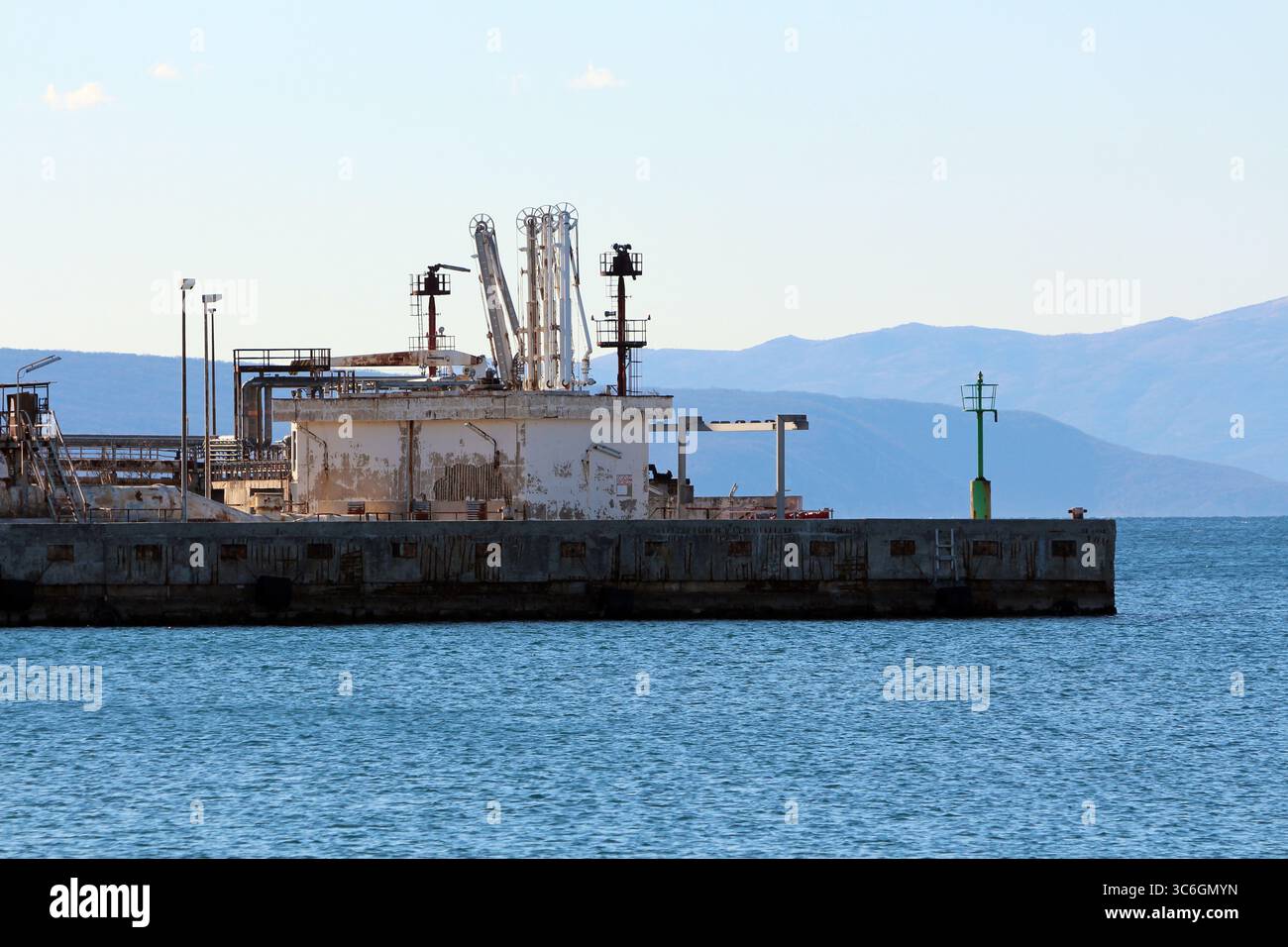 Un vecchio terminal marino sul molo presenta attrezzature arrugginite e strutture industriali adagiate lungo l'acqua blu, incorniciate da montagne lontane sotto un pallido Foto Stock